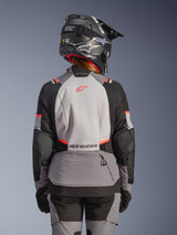 Woman rider from behind wearing the Stella Andes V4 Drystar® Jacket in Dark Gray Ice Gray Black Coral Fluo, paired with matching adventure pants and a black helmet, featuring a large light gray back panel with coral accents and Alpinestars branding.