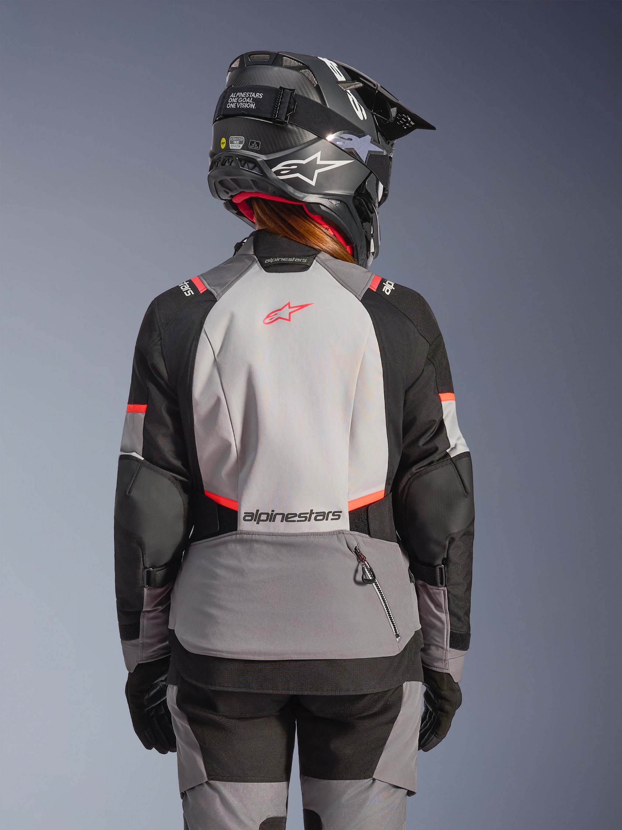 Woman rider from behind wearing the Stella Andes V4 Drystar® Jacket in Dark Gray Ice Gray Black Coral Fluo, paired with matching adventure pants and a black helmet, featuring a large light gray back panel with coral accents and Alpinestars branding.