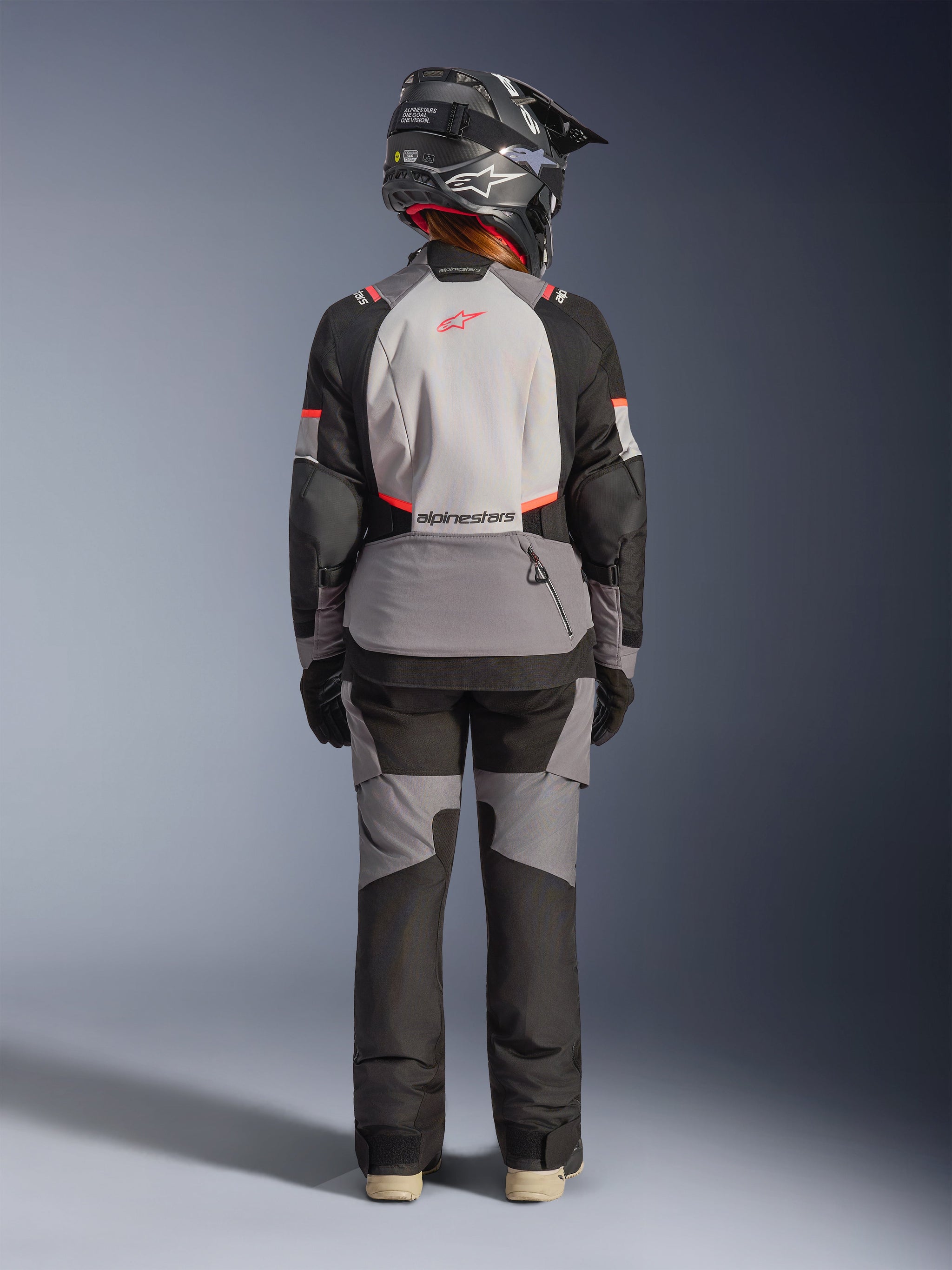 Woman wearing Alpinestars Stella Andes V4 Drystar® Jacket from the back, Dark Gray, Ice Gray, Black, and Coral Fluo accents, matching adventure pants and black helmet, multi-paneled textile design for touring and all-weather performance