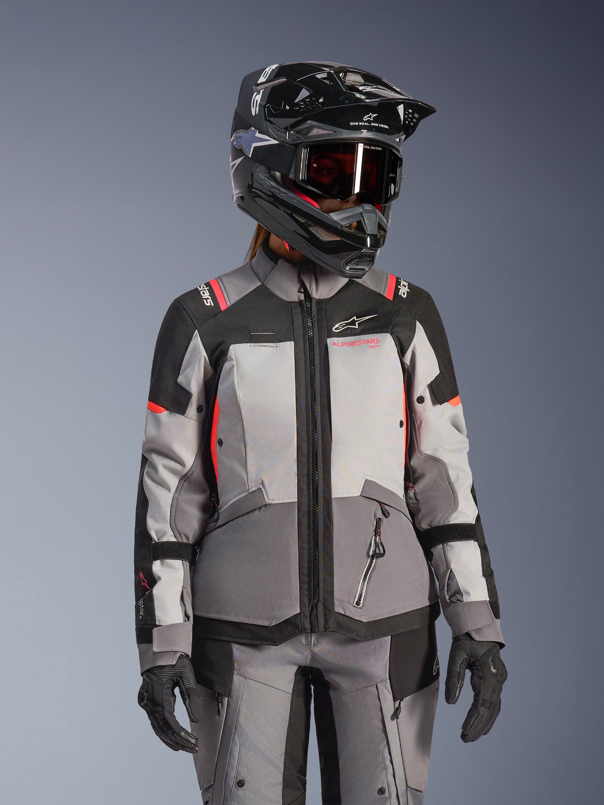 Woman Stella Andes V4 Drystar® Jacket, Motorcycle Touring Jacket, Dark Gray and Ice Gray with Black and Coral Fluo accents, worn with a black helmet and goggles, technical waterproof gear for adventure riding.