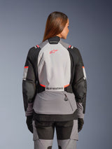 Woman wearing the Alpinestars Stella Andes V4 Drystar® Jacket, rear view, Dark Gray Ice Gray Black and Coral Fluo, features high-visibility coral accents and branding on the back, technical textile construction for adventure motorcycling