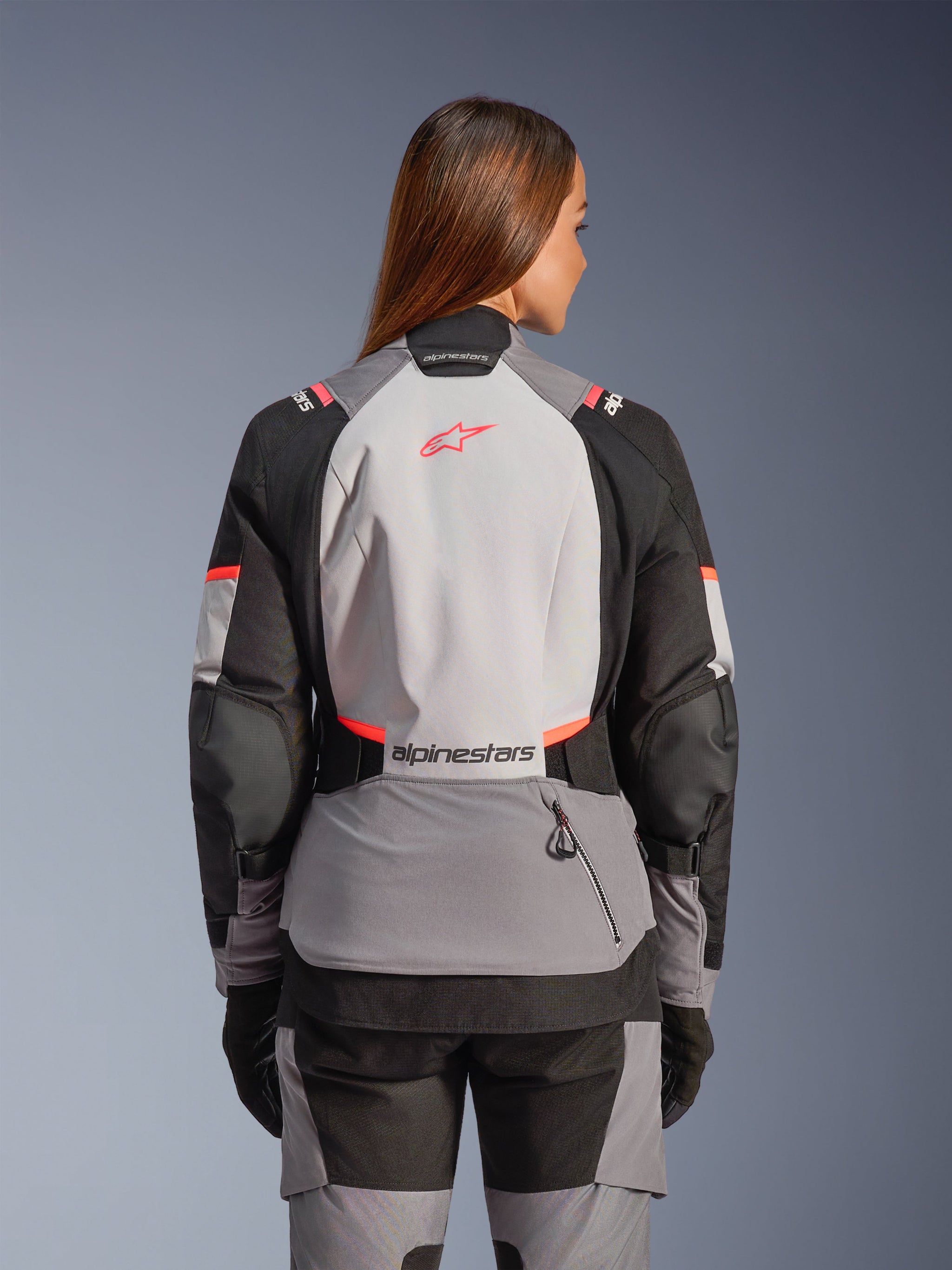 Woman wearing the Alpinestars Stella Andes V4 Drystar® Jacket, rear view, Dark Gray Ice Gray Black and Coral Fluo, features high-visibility coral accents and branding on the back, technical textile construction for adventure motorcycling