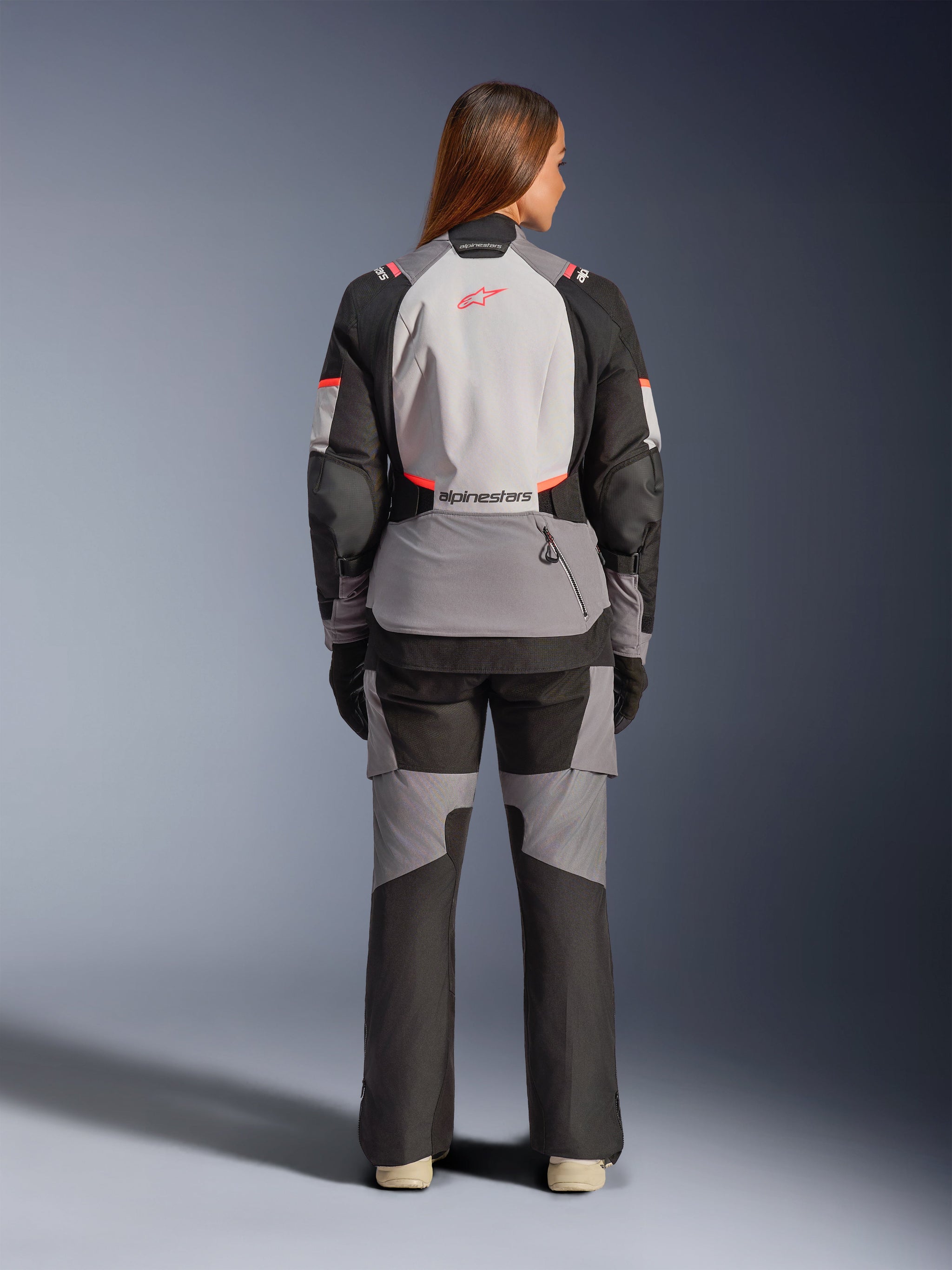 Female rider from behind wearing the Alpinestars Stella Andes V4 Drystar® Jacket and matching pants, Dark Gray Ice Gray Black and Coral Fluo, adventure touring motorcycle gear with Alpinestars logo on the back, all-weather protective apparel.