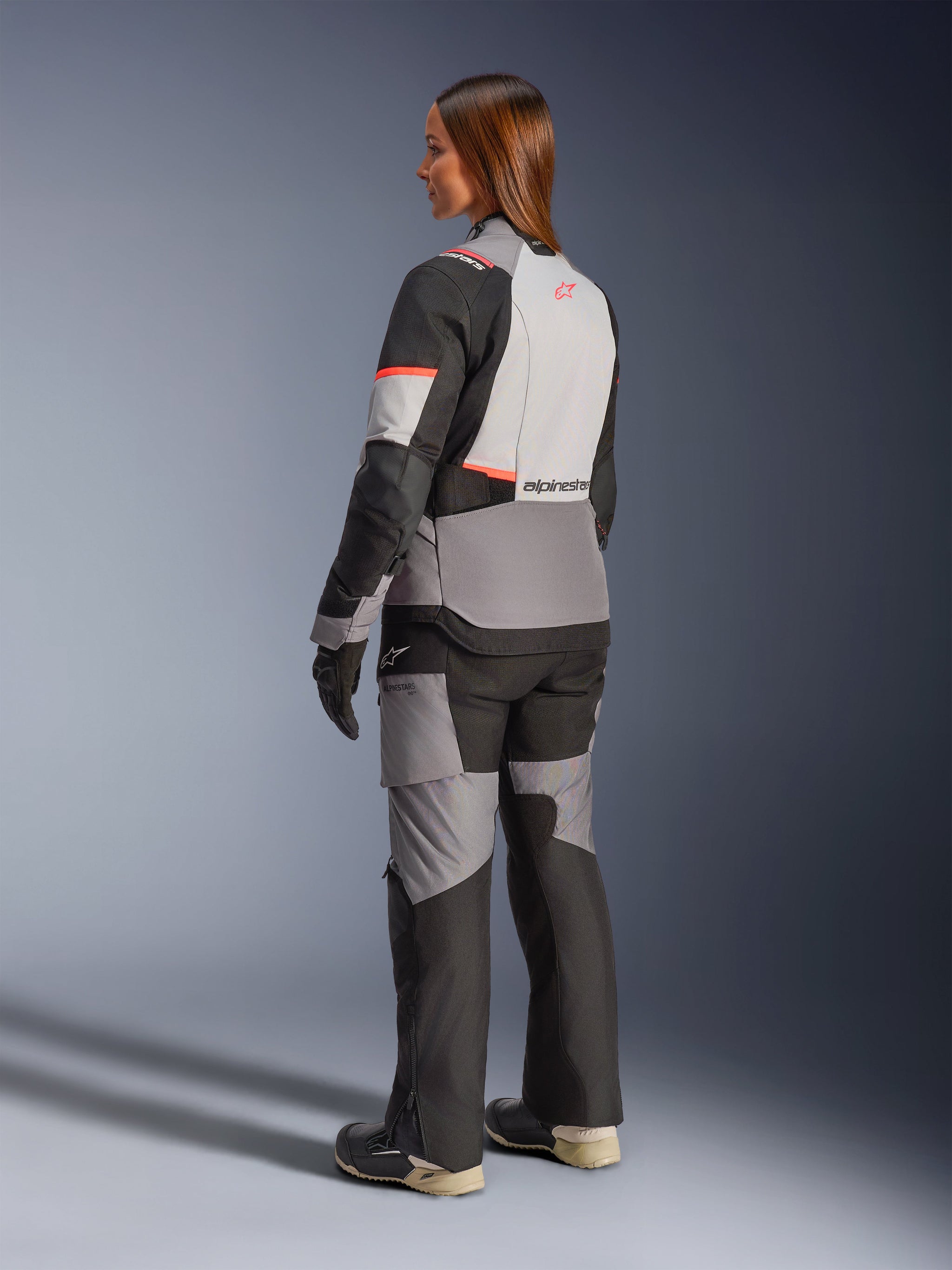 Woman wearing the Alpinestars Stella Andes V4 Drystar® Jacket in Dark Gray, Ice Gray, Black, and Coral Fluo, rear view, paired with matching adventure motorcycle pants and boots, showcasing the sleek modern design and high-visibility accents.