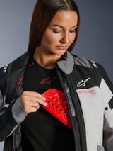 Woman wearing the Alpinestars Stella Andes V4 Drystar® Jacket in Dark Gray and Ice Gray, inserting a red protective chest armor into the internal compartment, demonstrating safety features and durable textile construction
