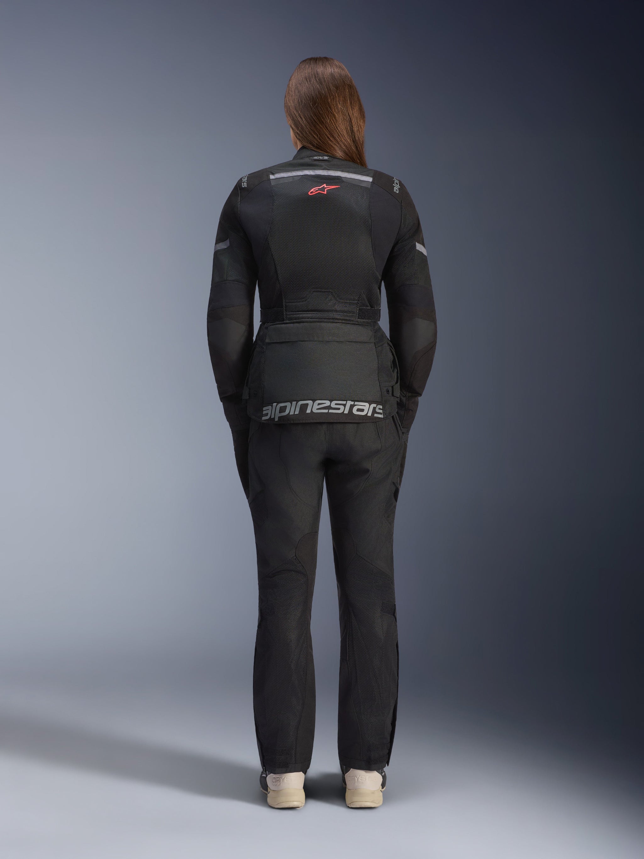 Woman wearing Alpinestars Stella Andes Air Drystar® Jacket and matching pants, Black, back view showing large grey alpinestars branding on the lower back pouch, small red star logo on upper back, mesh ventilation panels, and reflective details for adventure touring