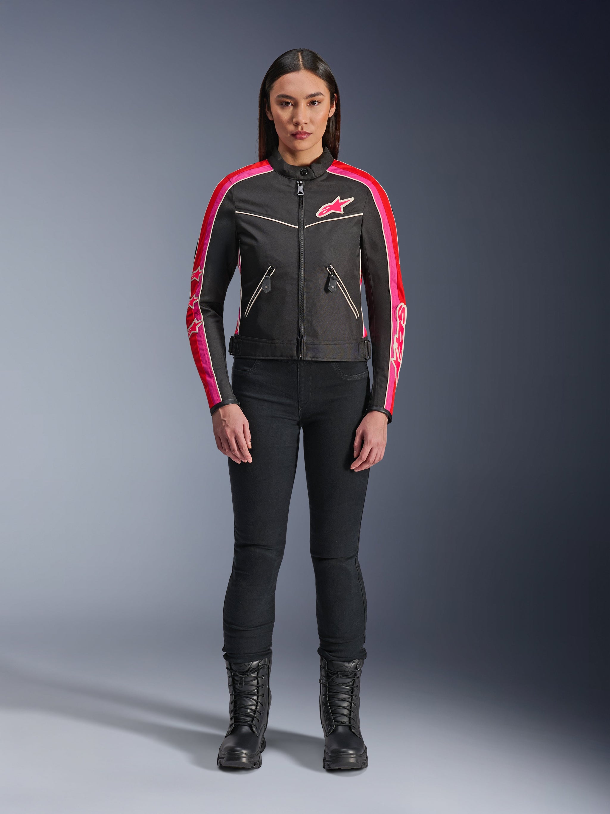 Woman wearing Alpinestars Stella T-Dyno WR Jacket, Black with Bright Red and Fuchsia accents, vintage-style textile jacket with pink logo and sleeve stripes, worn with black pants and lace-up boots, urban motorcycle gear