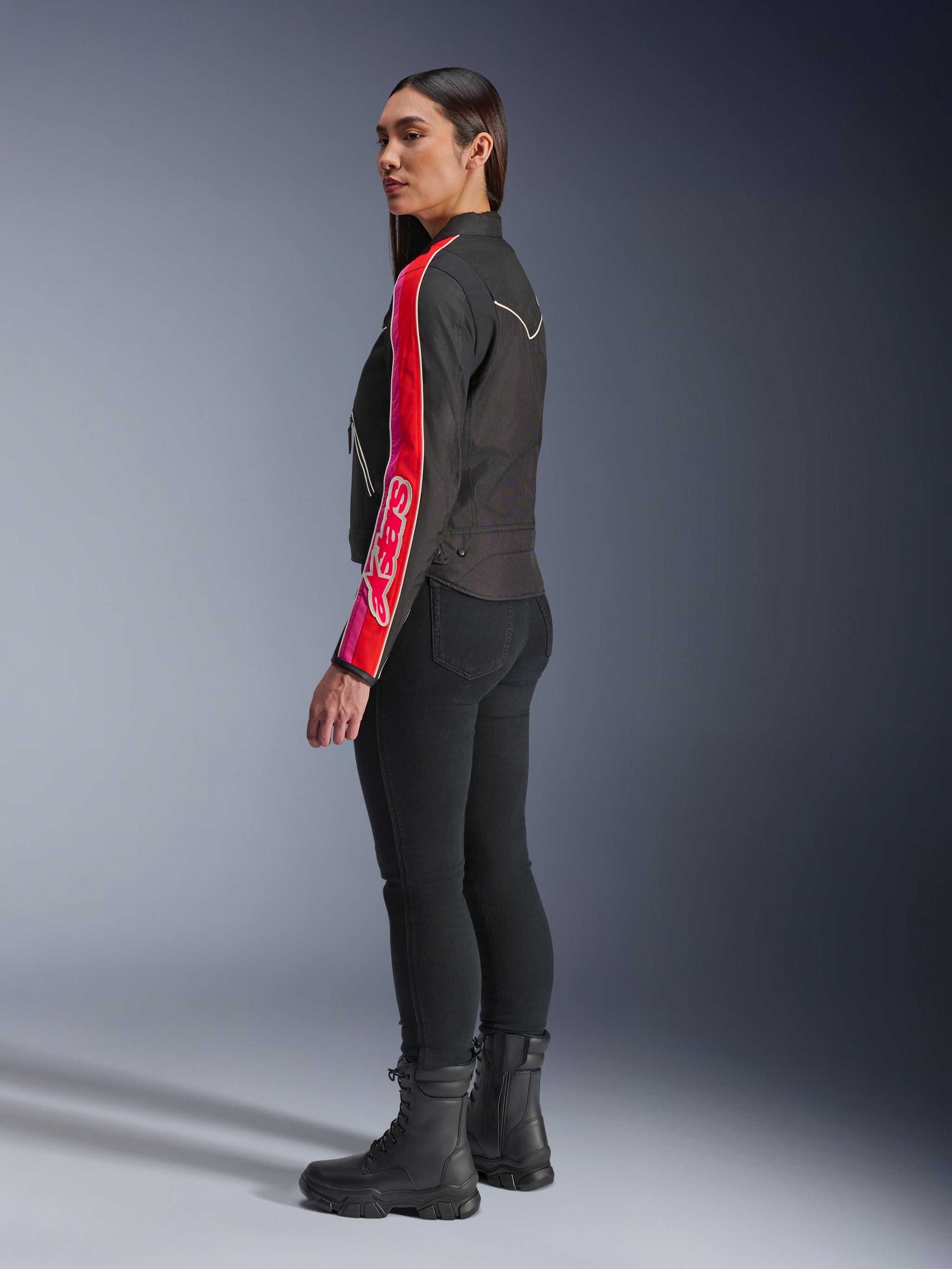 Female model wearing the Alpinestars Stella T-Dyno WR Jacket in Black Bright Red Fuchsia, featuring bold red and pink stripes with Alpinestars branding on the sleeve, paired with black riding pants and black motorcycle boots in a studio setting.