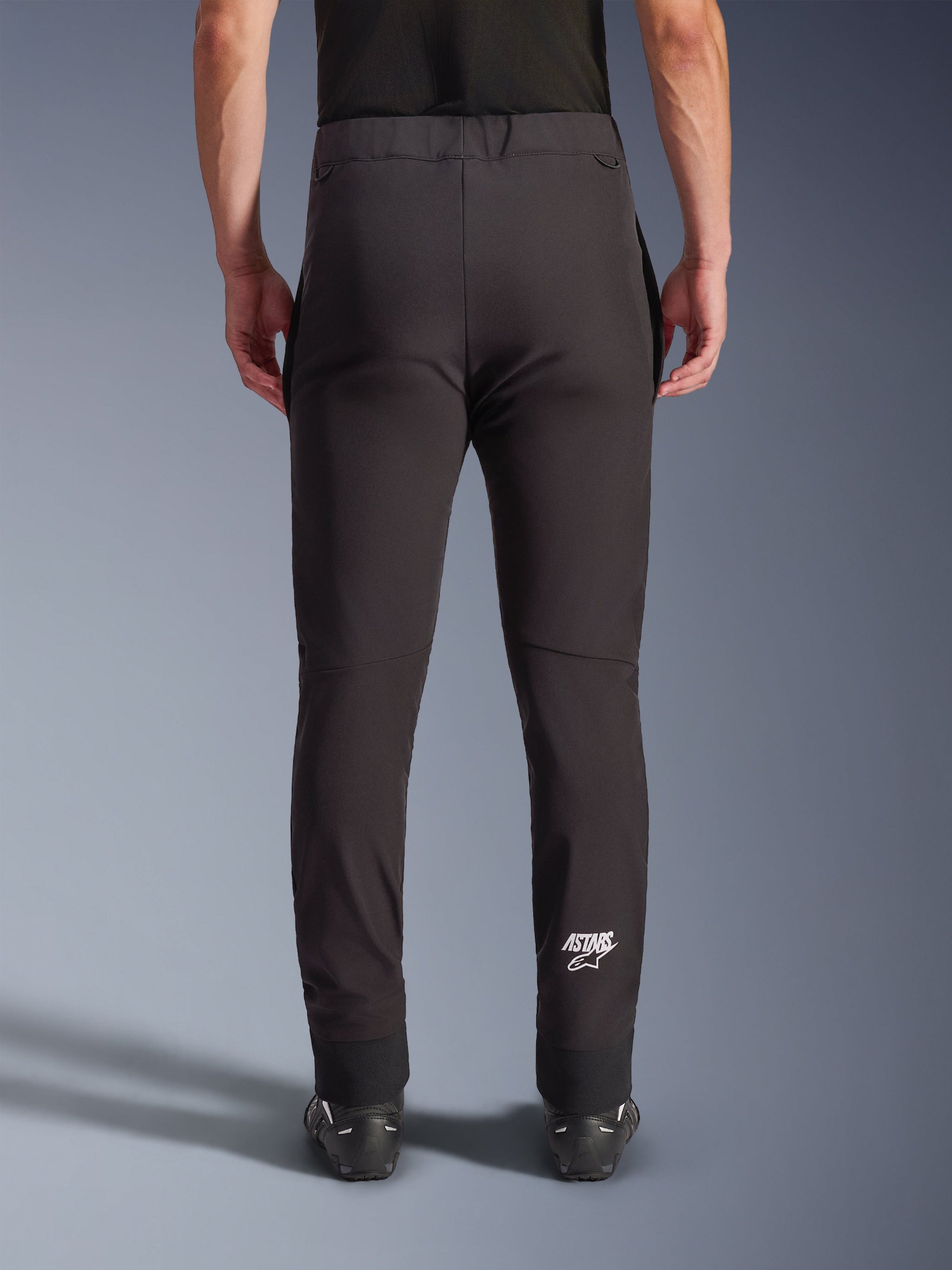 Alpinestars Aeroshell Pants, Black softshell motorcycle pants, back view showing slim fit and pre-curved knees, white Astars logo on the lower right calf, elasticated lower hem, crossover design for road riding