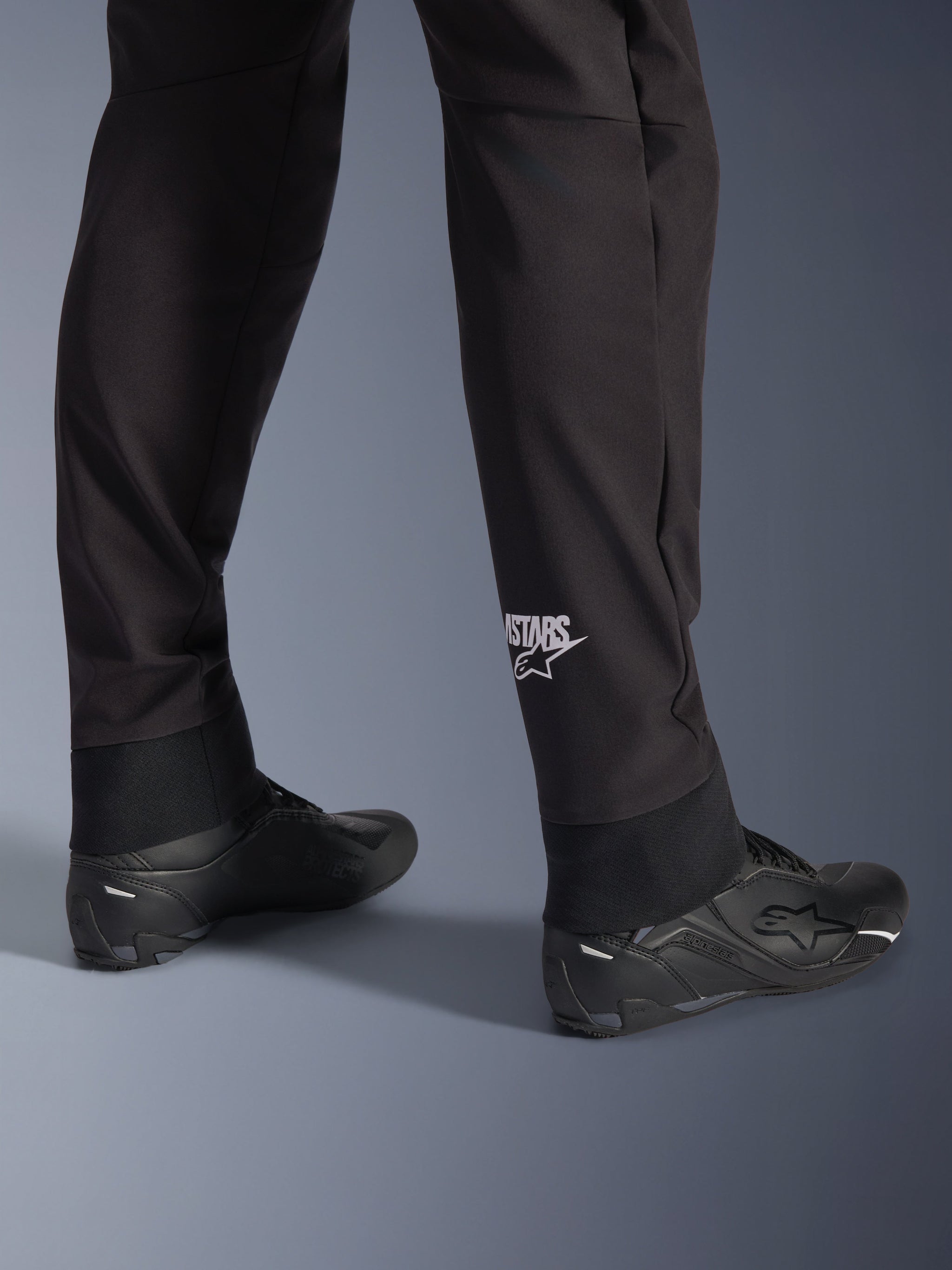 Alpinestars Aeroshell Pants, Black softshell motorcycle pants, close-up of lower leg and elasticated hem, white Alpinestars logo on calf, styled with black riding shoes, modern crossover sport design