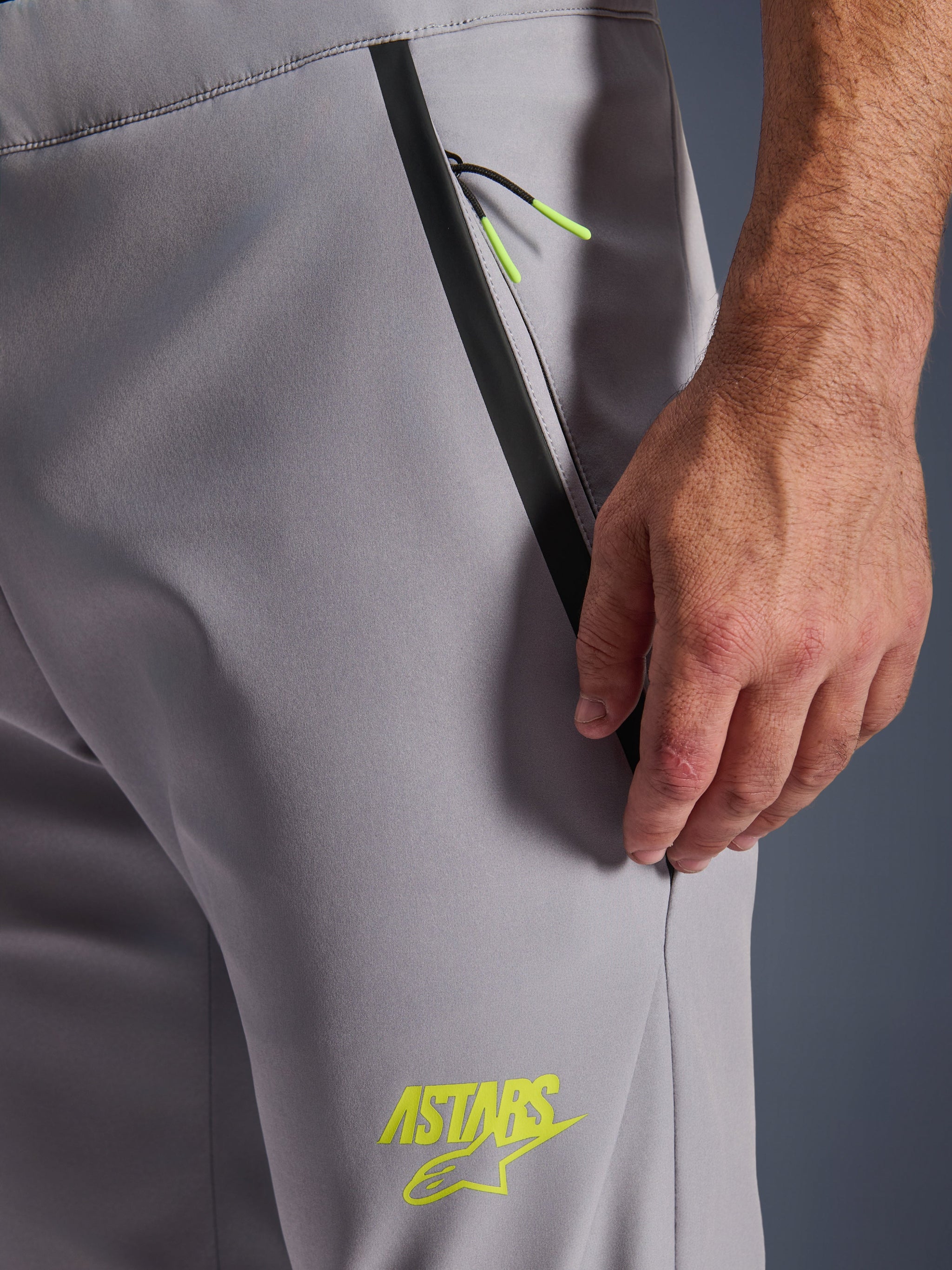 Alpinestars Aeroshell Pants, Mid Gray with Yellow Fluo accents, close-up view of the zipped front pocket with black taped trim and bright yellow pull cords, yellow ASTARS logo on the thigh, softshell stretch material for motorcycle riding.