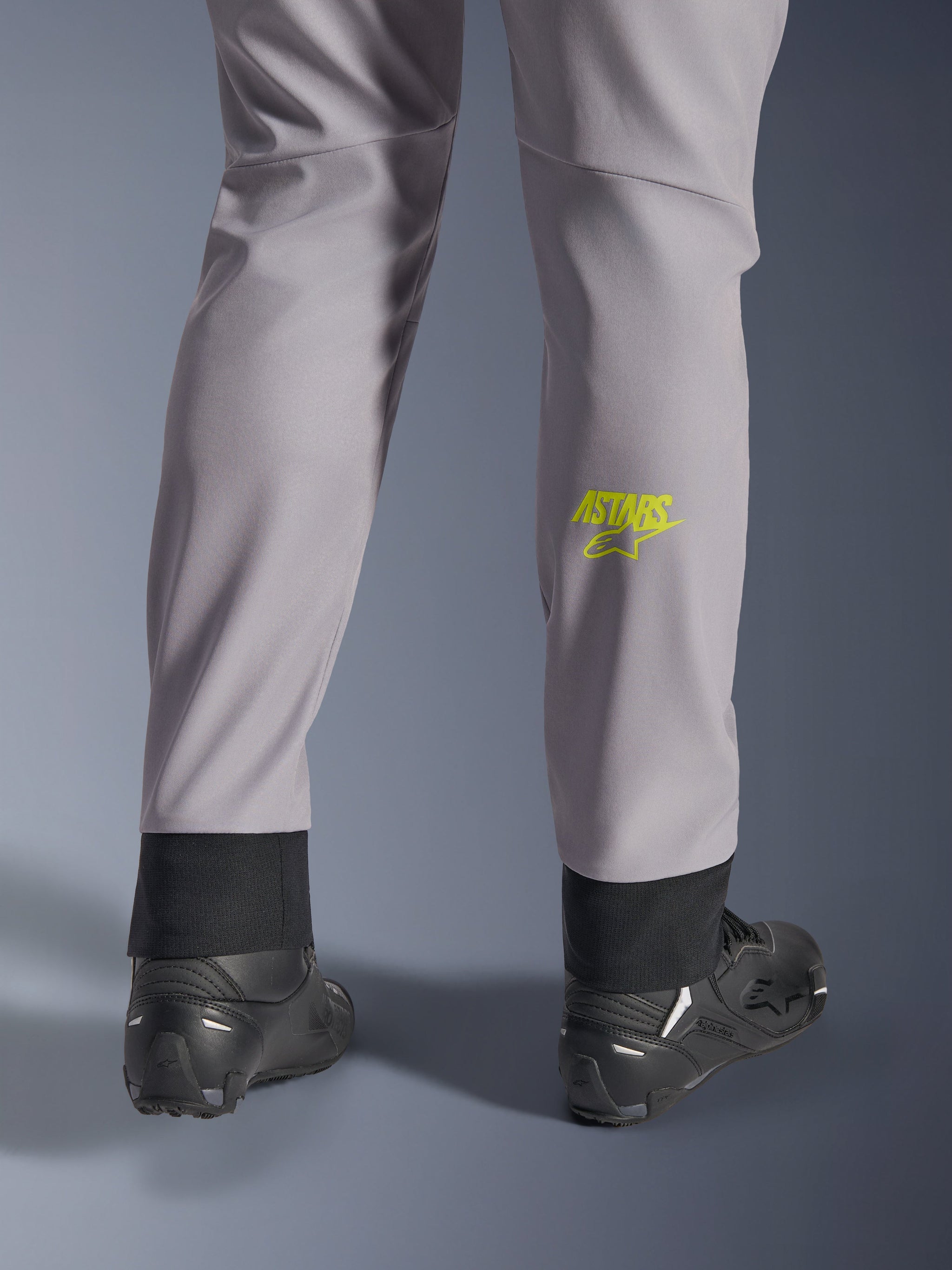 Alpinestars Aeroshell Pants, Mid Gray with Yellow Fluo Astars logo on the calf, shown from the back wearing black motorcycle shoes, featuring an elastic lower hem and a modern tapered silhouette