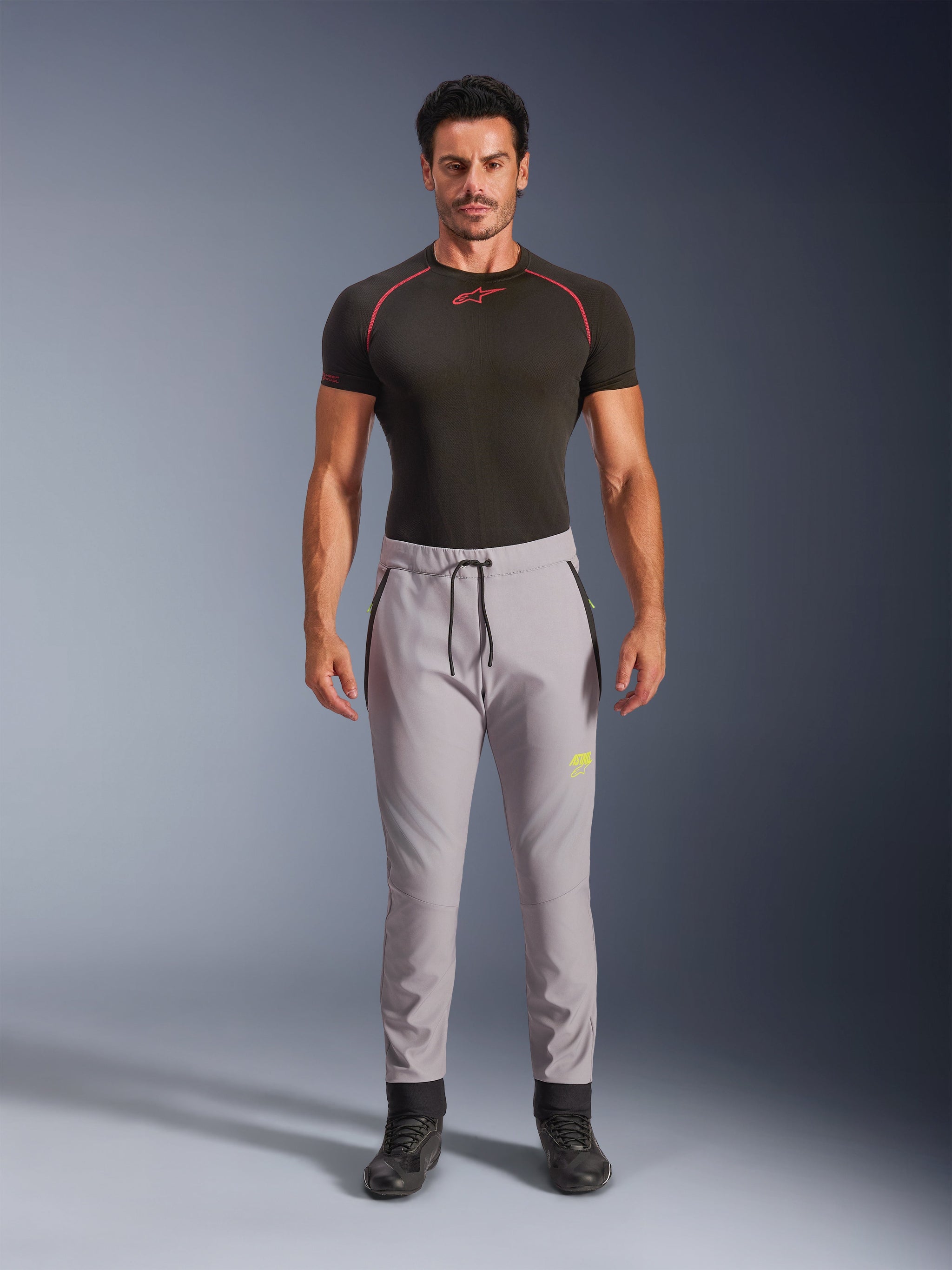 Male model wearing Alpinestars Aeroshell Pants in Mid Gray and Yellow Fluo, crossover sports softshell pants with elastic drawstring waist and zippered pockets, paired with a black short-sleeved performance shirt, modern athletic design for road and casual riding