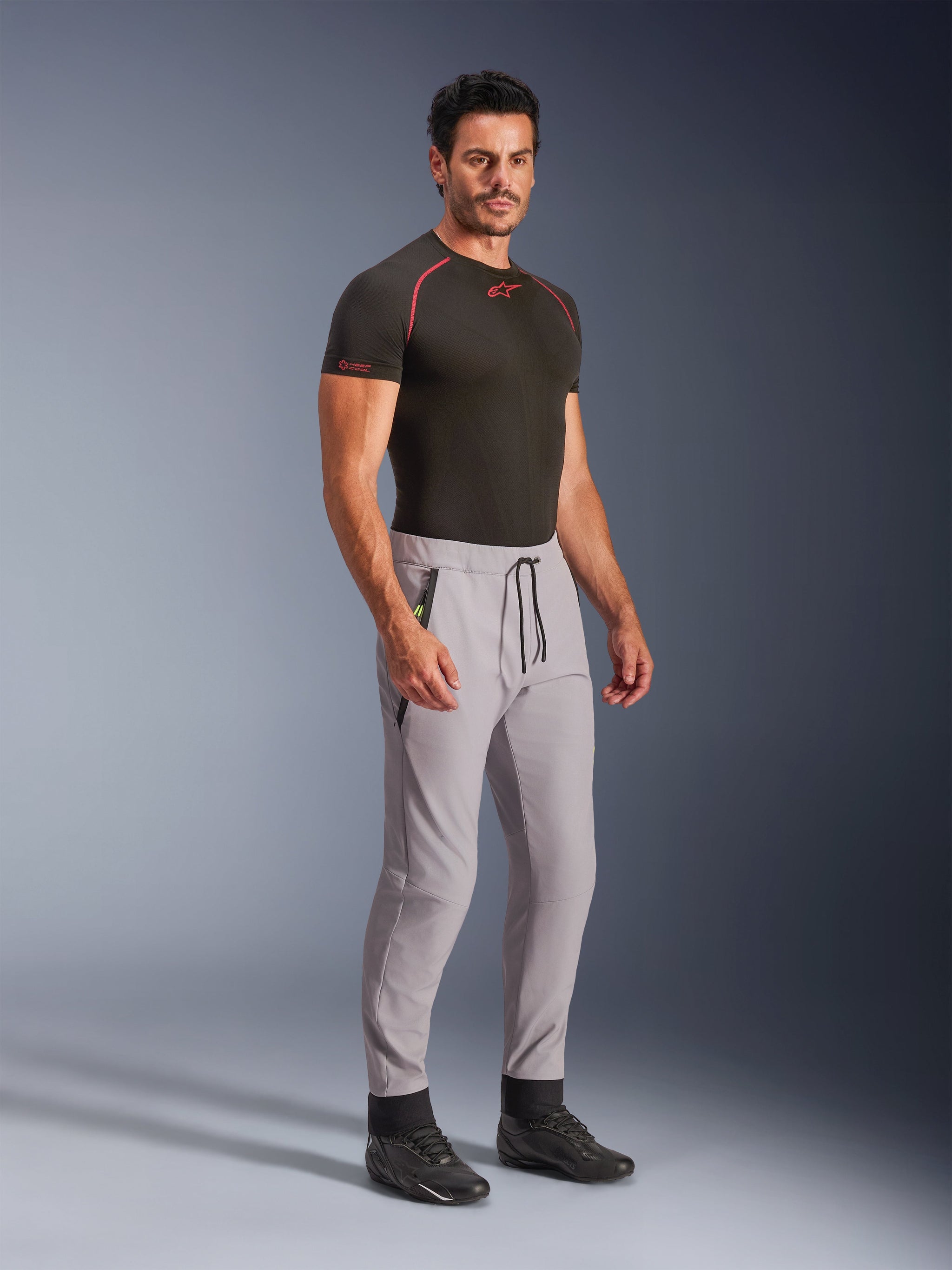 Alpinestars Aeroshell Pants, Mid Gray with Yellow Fluo accents, crossover softshell pants worn by a model with a black base layer and black shoes, featuring a modern tapered fit, elastic waistband with black drawcord, and zipped pockets.