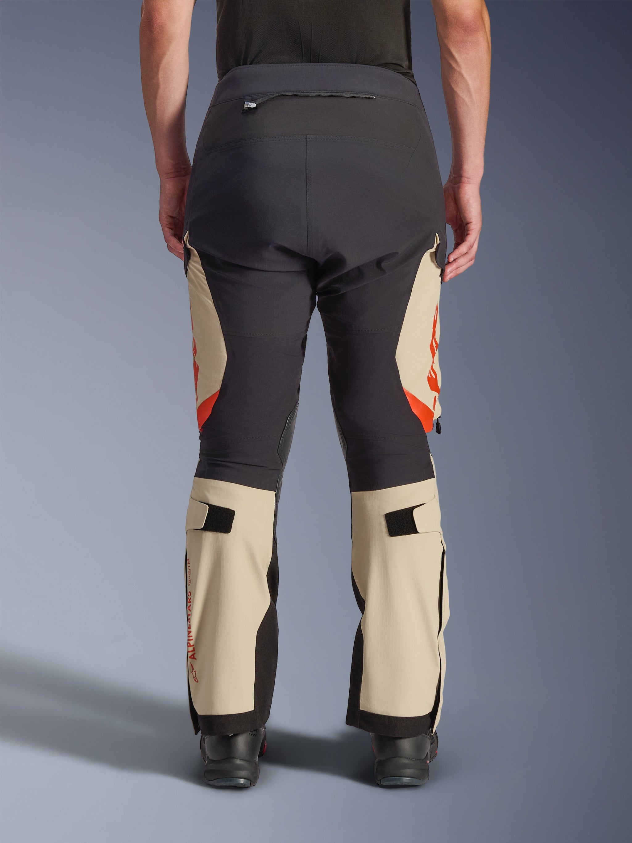Rear view of Alpinestars Halo Pro Drystar® XF Laminated Pants, Mastic Black with tan panels and orange accents, adventure motorcycle pants featuring a technical textile construction, worn with black riding boots
