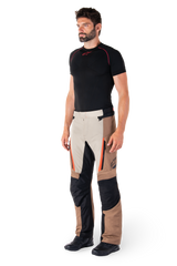 Man wearing Alpinestars ST-1 Waterproof Pants, Adventure Touring Pants, Dark Khaki Sand Black and Red Fluo, styled with a black base layer shirt and black motorcycle boots, featuring reinforced panels and orange zipper accents