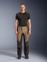 Male model wearing Alpinestars Andes V4 Drystar® Pants in Walnut Black, adventure motorcycle pants paired with a black base layer and boots, featuring orange zipper accents on the thighs and reinforced black panels for all-terrain durability.