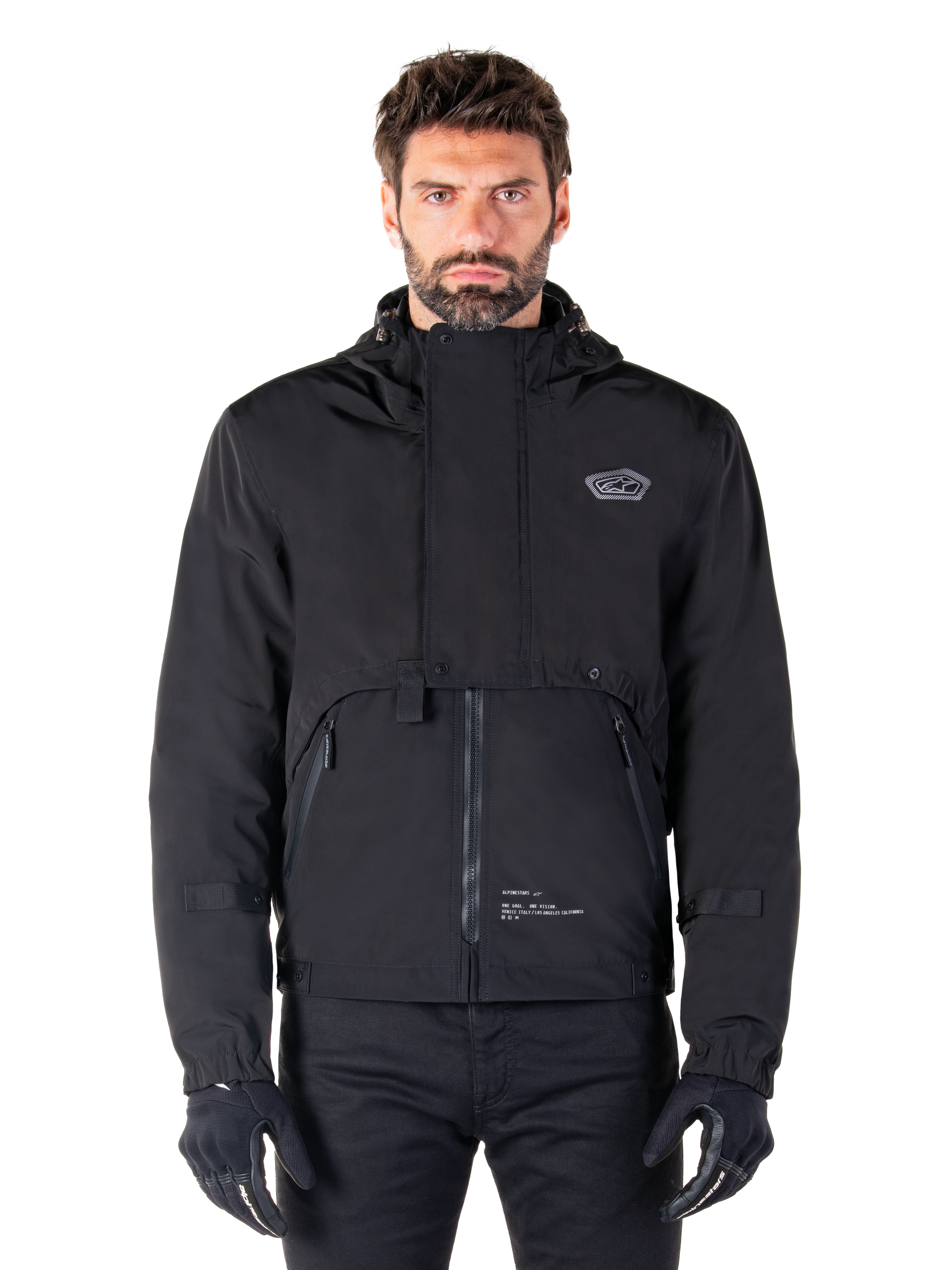 Alpinestars Mootant Waterproof Parka, Black hooded motorcycle jacket with a modular technical design, worn by a male model with black gloves, featuring a unique chest flap and zippered side pockets, urban and touring riding style.