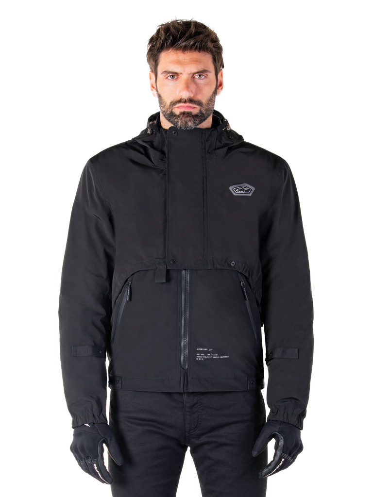 Alpinestars Mootant Waterproof Parka, Black hooded motorcycle jacket with a modular technical design, worn by a male model with black gloves, featuring a unique chest flap and zippered side pockets, urban and touring riding style.