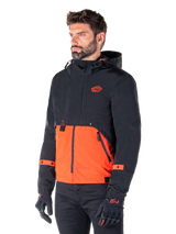 Man wearing the Alpinestars Mootant Waterproof Parka in Spicy Orange and Black, a modular motorcycle jacket with a black hooded upper layer and bright orange lower section, styled with black riding gloves and pants.