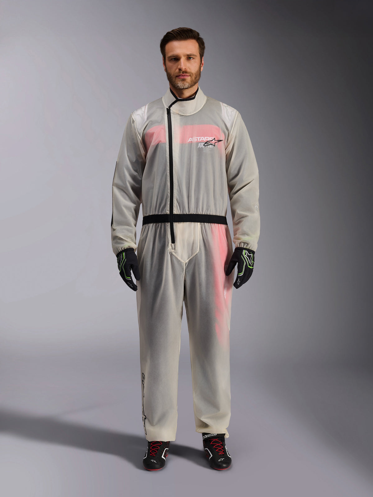 Alpinestars Kart Rain Suit, Clear, transparent waterproof oversuit worn by a model, featuring a black asymmetric zipper and chest logo, red and white racing suit visible underneath, paired with black and neon green gloves and black racing boots.