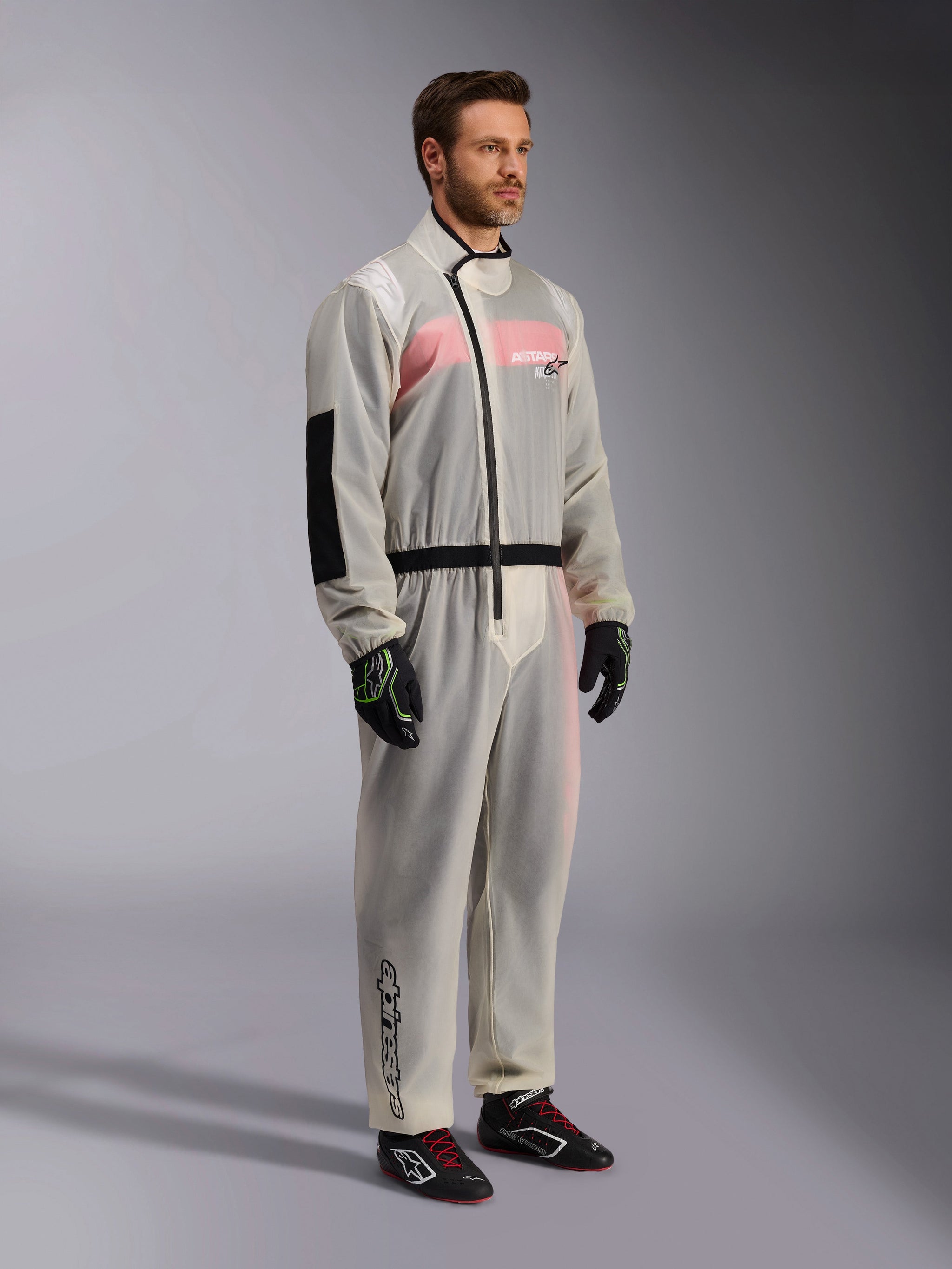 Alpinestars Kart Rain Suit, Clear waterproof karting oversuit, semi-transparent material showing a pink and white racing suit underneath, featuring a black asymmetric zipper, reinforced elbow patch, and black waist band, worn with black karting gloves and racing shoes.