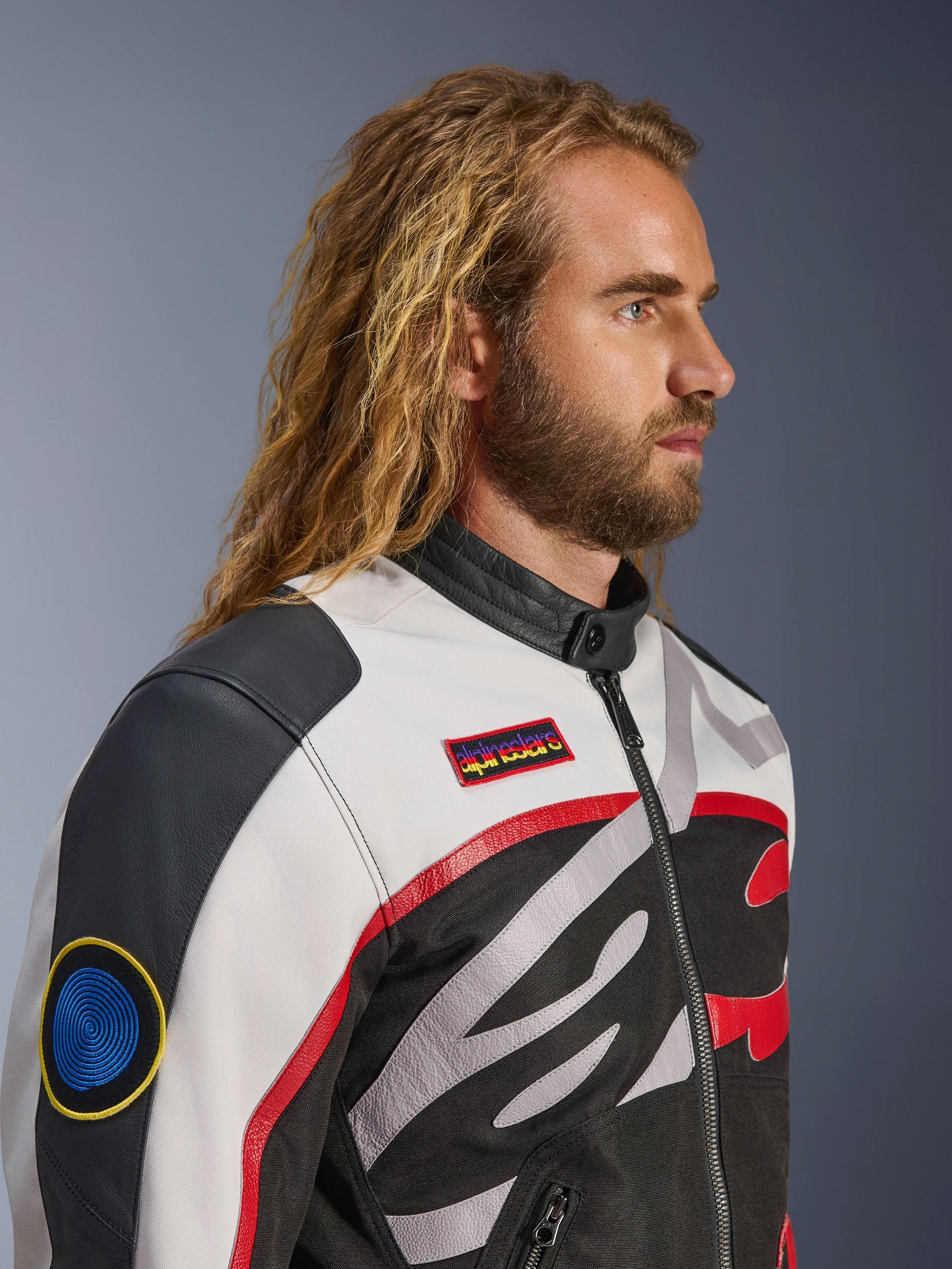 Man with long hair and beard wearing the Alpinestars Axton Jacket, Motorcycle Jacket, Black Off White and Bright Red, vintage racing-inspired design with branded patches and wrap-around logo, close-up profile view, urban contemporary style