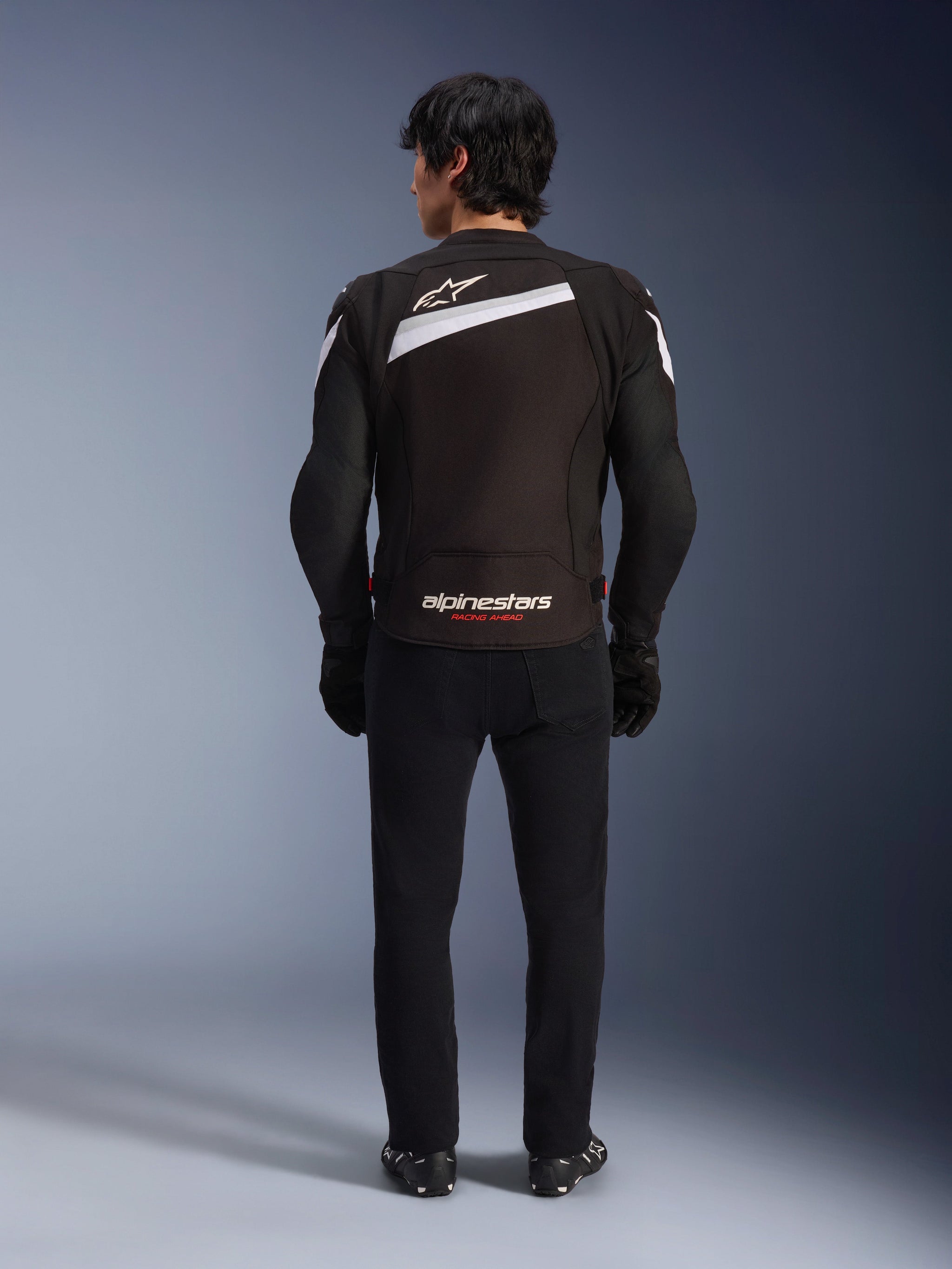 Alpinestars T-GP Plus R V4 Jacket, Black and White sport textile motorcycle jacket, rear view featuring white Alpinestars logo and 'RACING AHEAD' branding, worn with black trousers and boots, streamlined design for performance riding.