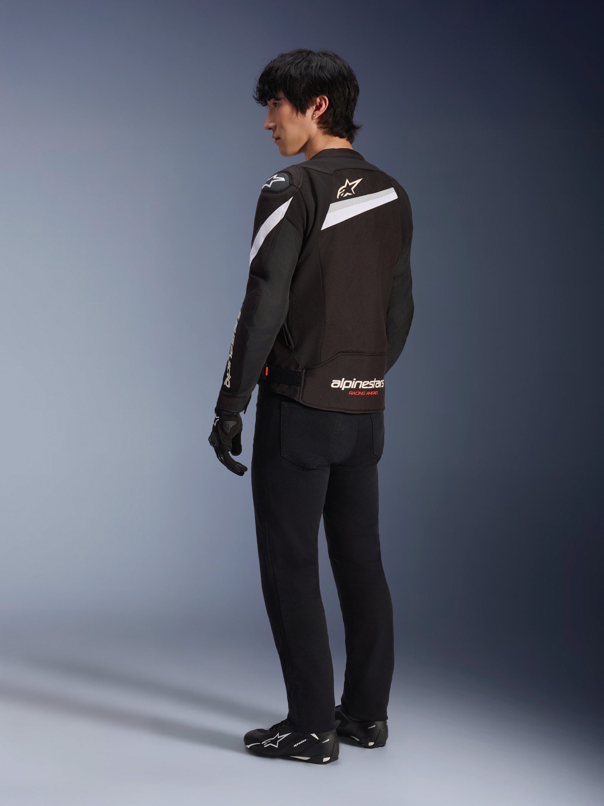Male rider wearing the Alpinestars T-GP Plus R V4 Jacket in Black and White, rear view showing white diagonal accents and branding on the lower back, paired with black riding pants, gloves, and technical boots, sport-fit textile motorcycle gear