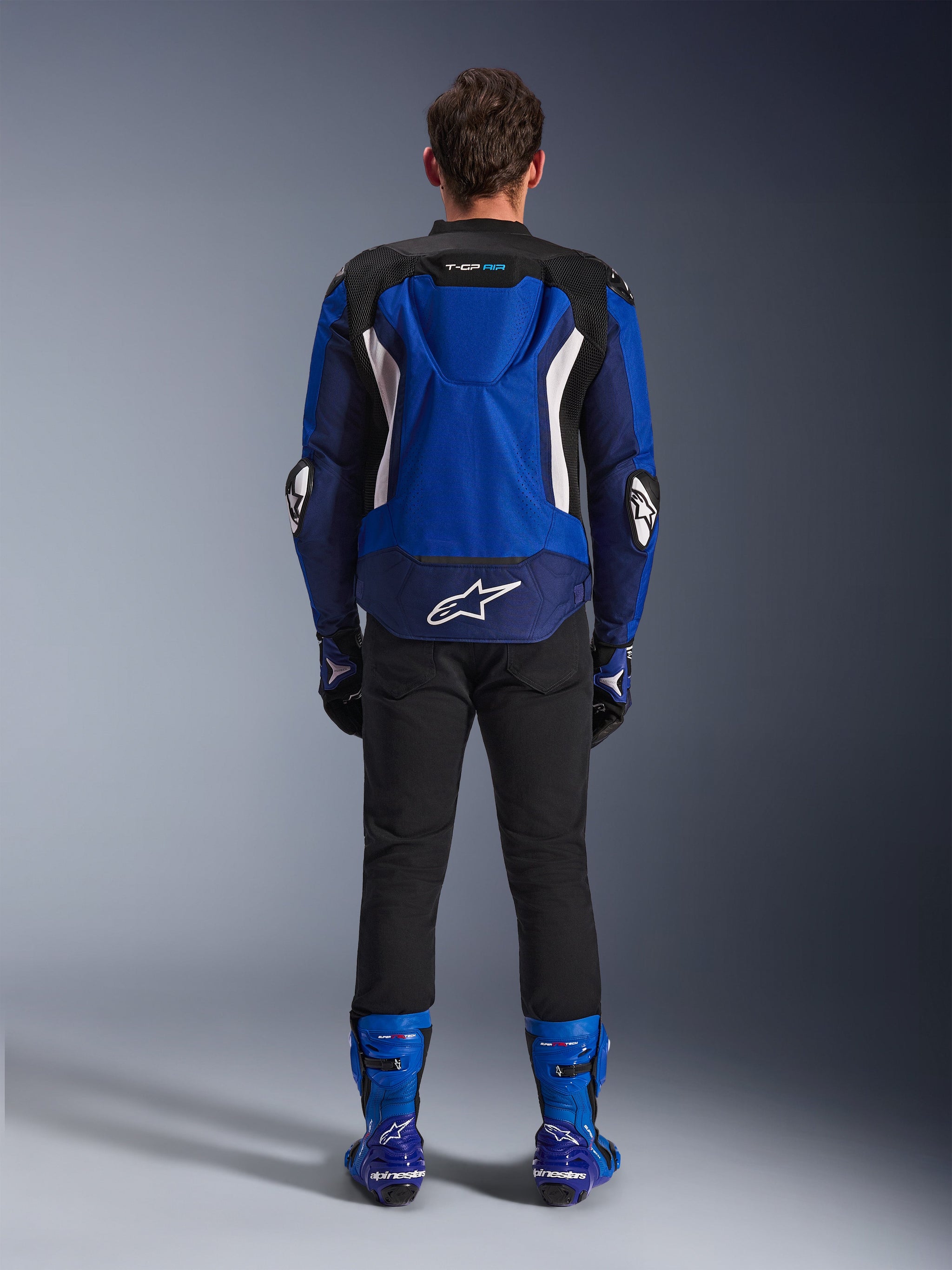 Alpinestars T-GP Air Jacket, Bright Blue Navy White, back view showing aerodynamic hump and mesh ventilation panels, white Alpinestars logo on lower back, worn with black pants and blue racing boots