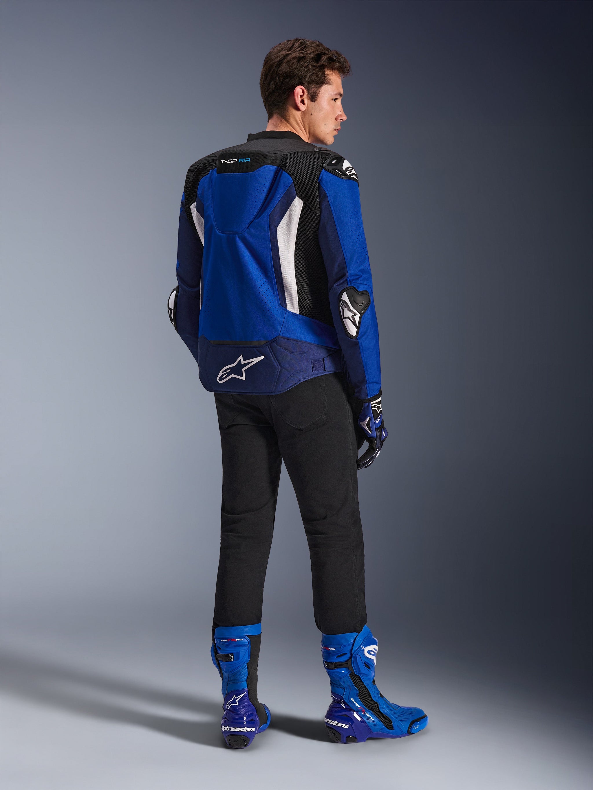 Alpinestars T-GP Air Jacket, Sport Textile Jacket, Bright Blue Navy White, viewed from behind on a model wearing black pants and matching blue and white boots, featuring white Alpinestars logo on the lower back, mesh ventilation panels, and an aerodynamic hump