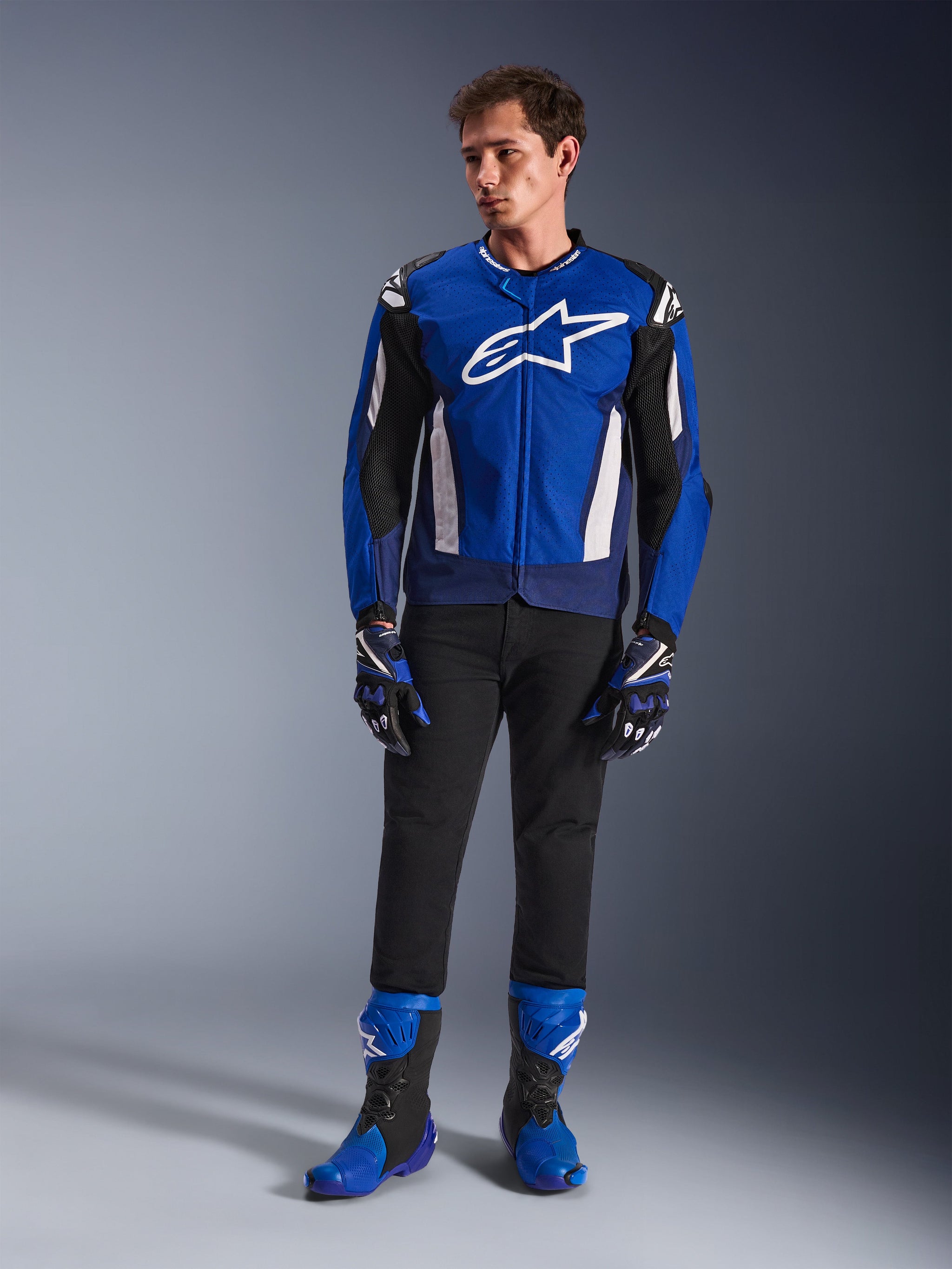 Male model wearing the Alpinestars T-GP Air Jacket in Bright Blue Navy White, paired with black pants, matching blue and navy racing gloves, and blue and black motorcycle boots, studio shot showcasing a complete sport riding outfit.