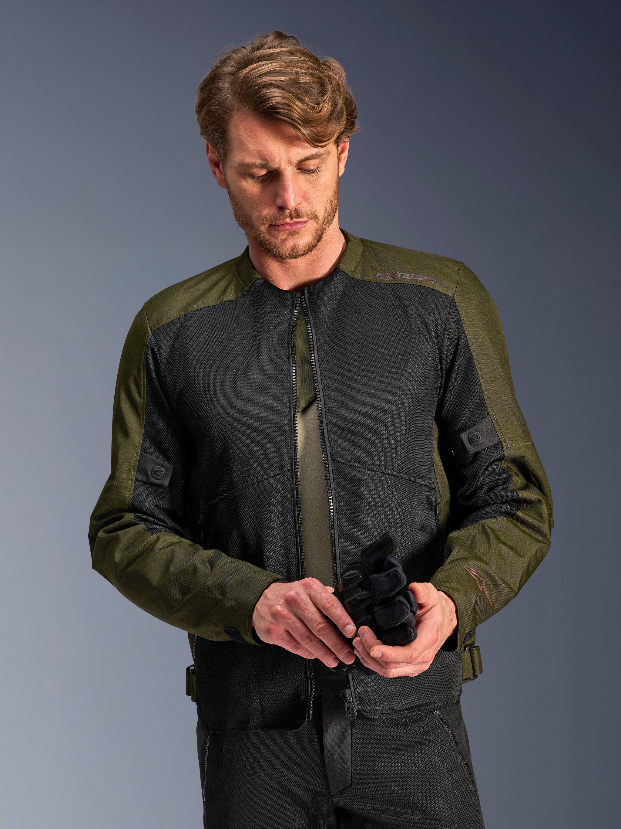 Man wearing the Alpinestars C-1 Air Jacket in Khaki, urban motorcycle jacket with olive green textile and black mesh panels, looking down while holding black motorcycle gloves, clean and contemporary style for road riding.