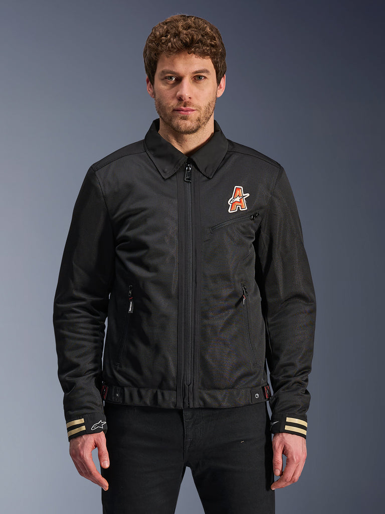 Alpinestars Ignite Air Jacket, Black motorcycle jacket, traditional shirt collar, orange and white 'A' logo on chest, beige striped cuffs with white star logo, urban contemporary riding gear