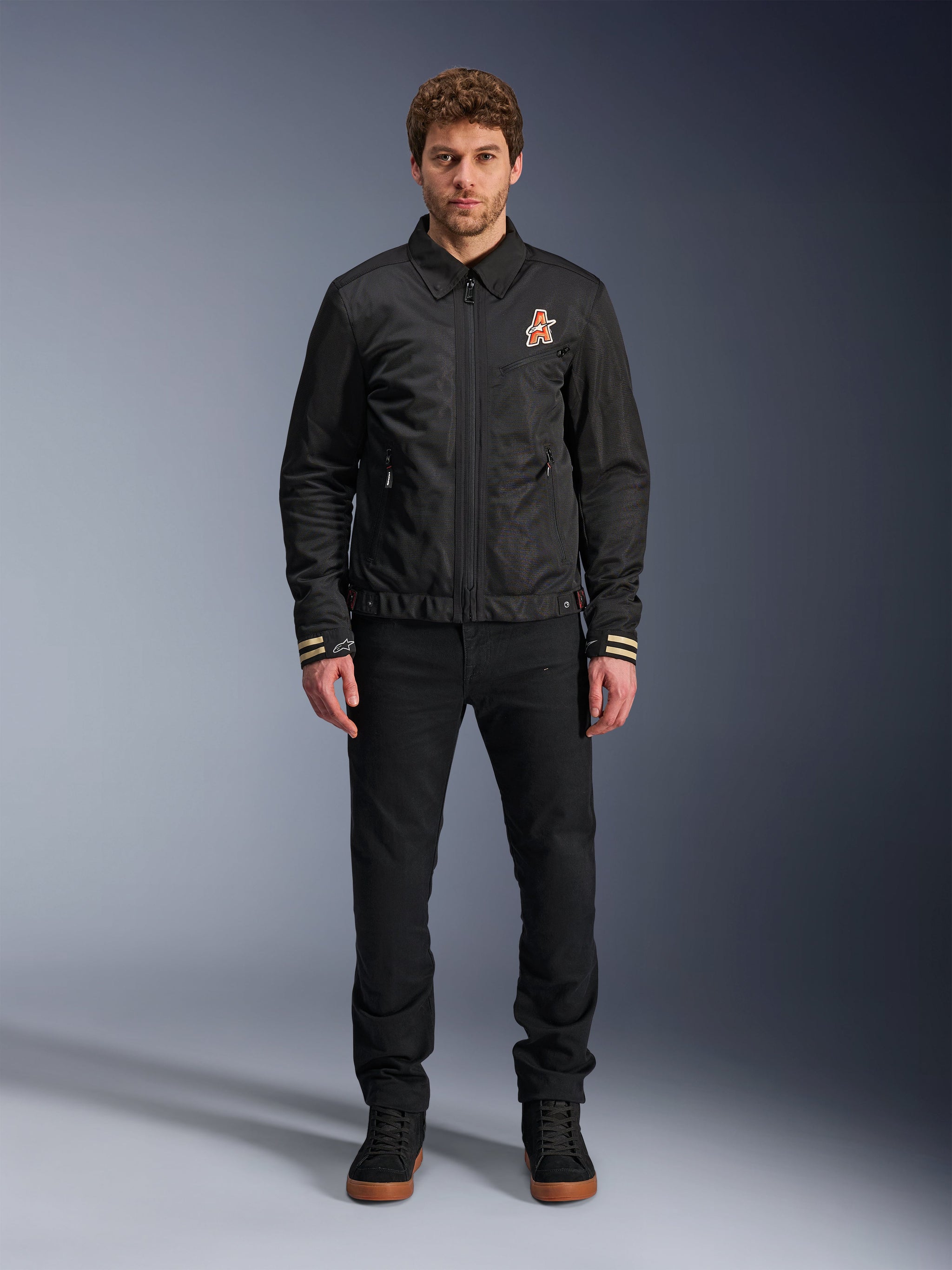 Male model wearing the Alpinestars Ignite Air Jacket in black, featuring a classic shirt collar, full front zipper, and an orange logo on the chest. Styled with black pants and black high-top sneakers for an urban contemporary motorcycle look.