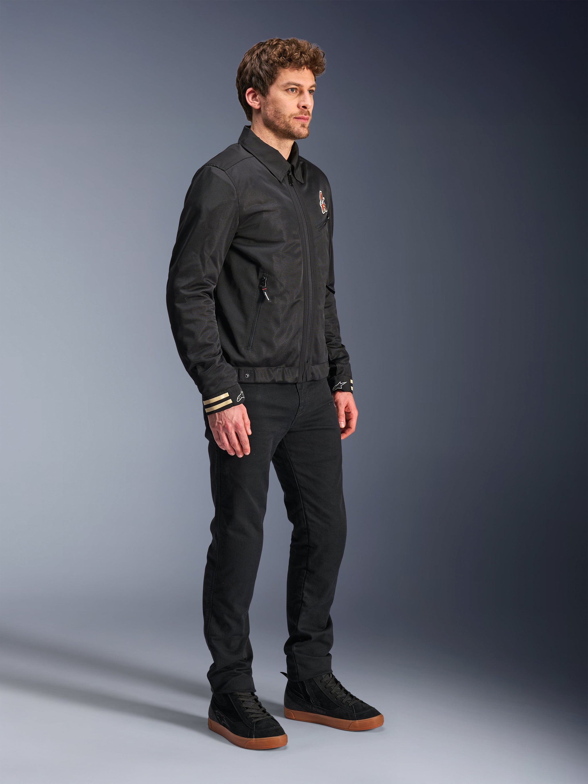 Alpinestars Ignite Air Jacket, Urban Motorcycle Jacket, Black, worn by a model with black pants and black sneakers, featuring a traditional shirt collar, club-inspired graphics on the chest and cuffs, sleek contemporary design with mesh and satin finish
