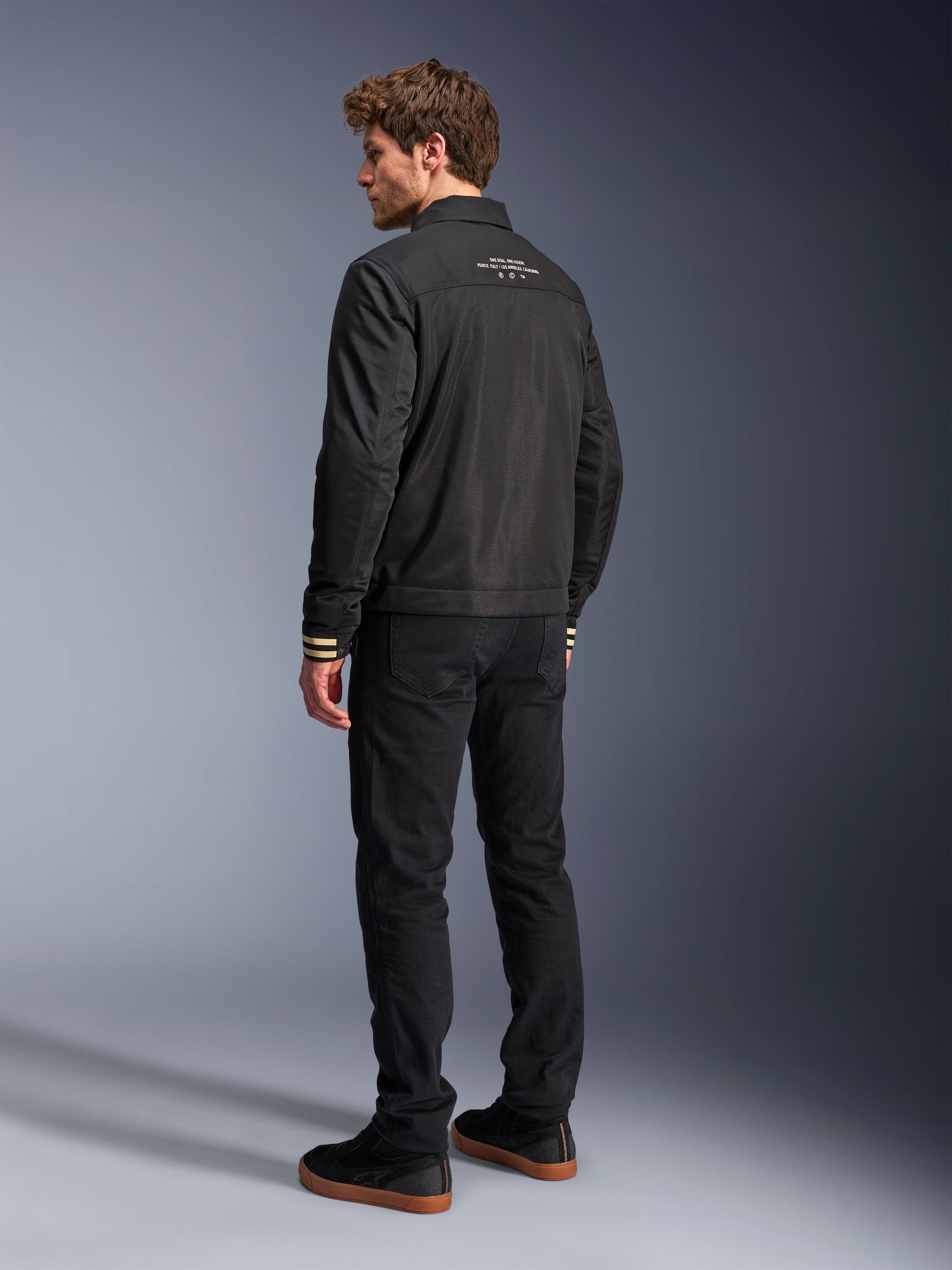 Alpinestars Ignite Air Jacket, Black motorcycle jacket, rear view featuring white graphic text on the upper back and tan striped cuffs, Harrington-style silhouette with mesh body, paired with black pants and black sneakers