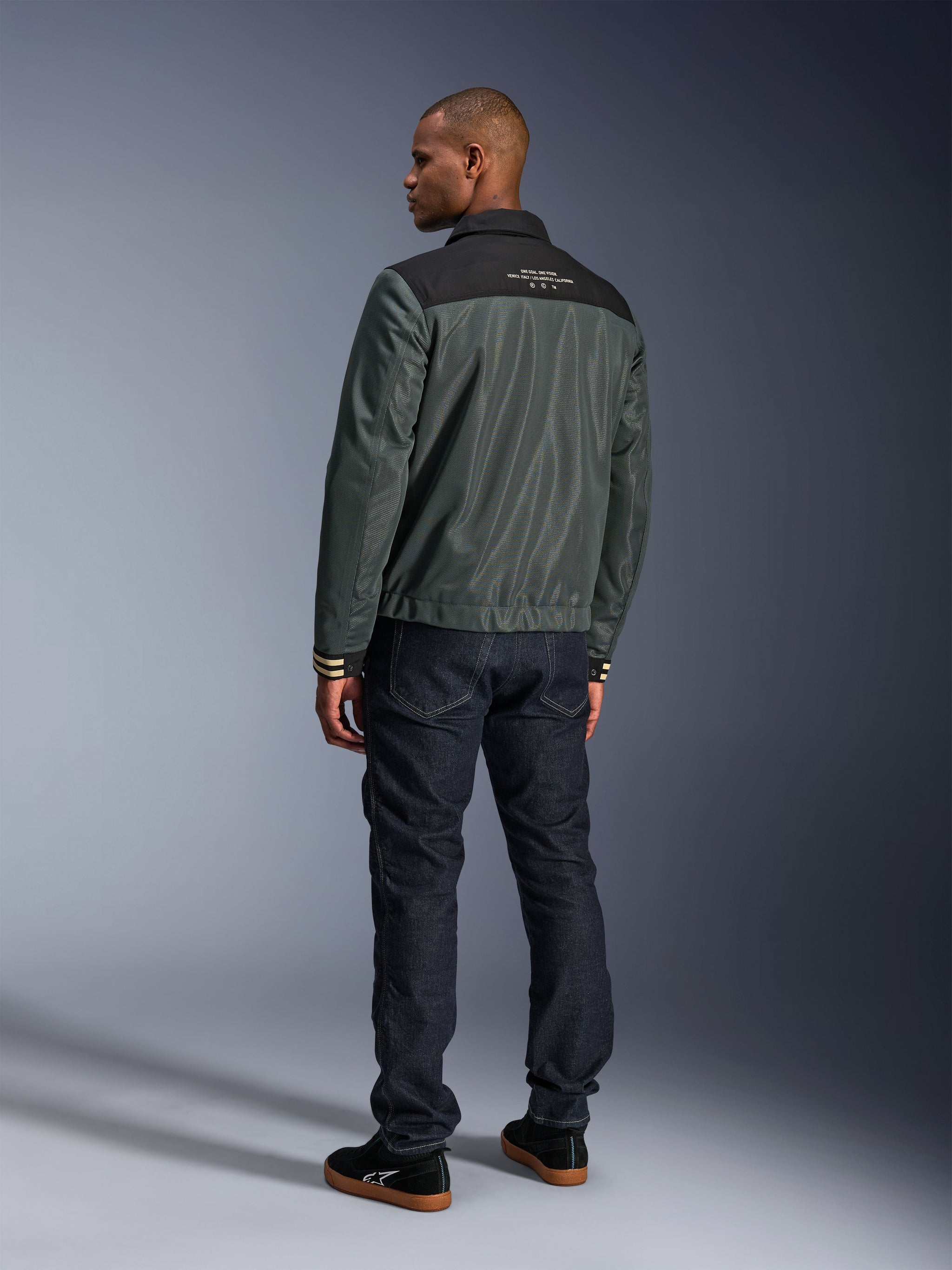 Alpinestars Ignite Air Jacket in Urban Gray Black Blue, rear view on model, featuring a mesh body with a black shoulder yoke and white text, striped cuffs, paired with dark denim jeans and black sneakers, urban lifestyle motorcycle apparel