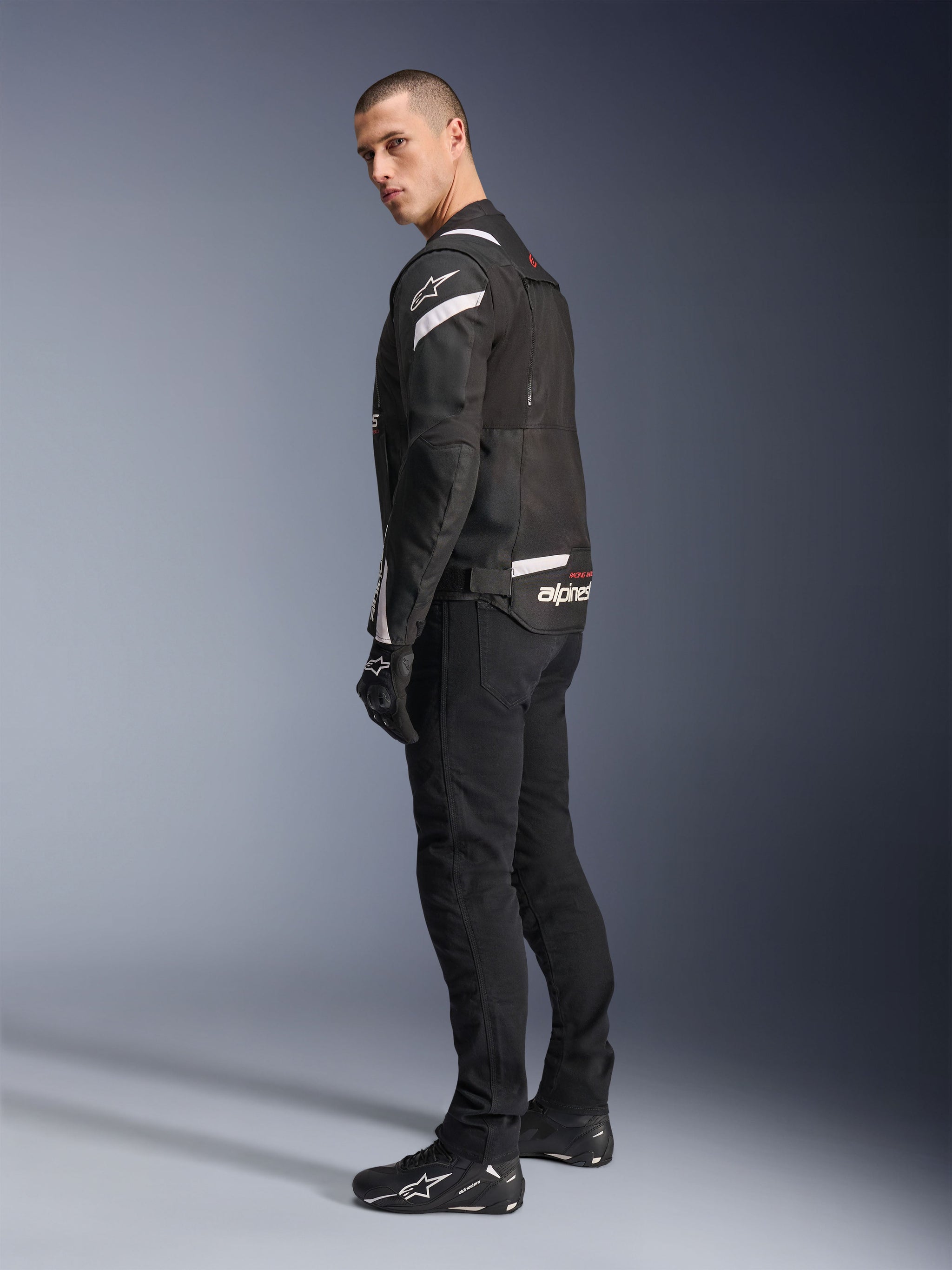 Alpinestars T-Stunt Air Jacket, Black and White, modular motorcycle jacket with tech mesh panels, shown from a back-side profile on a model, worn with black riding pants and shoes, featuring white accents and branding on the shoulders and back.