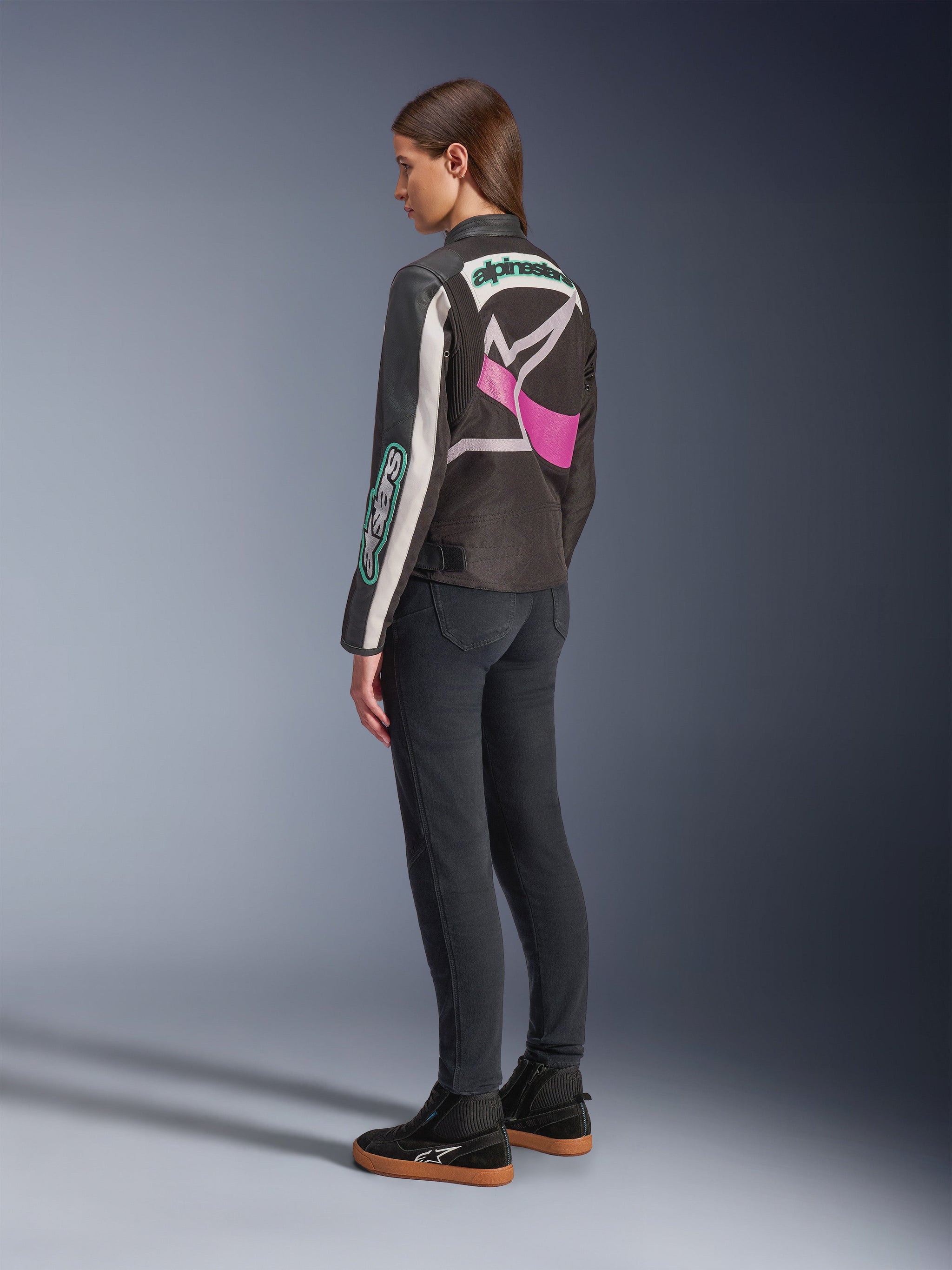 Alpinestars Idles Women’s Jacket, Motorcycle Jacket, Black Off White and Purple, viewed from the back on model, featuring a large wrap-around Alpinestars logo and vintage racing-inspired design, paired with dark jeans and black shoes.