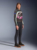 Female model wearing the Alpinestars Idles Women's Jacket, Motorcycle Jacket, Black Off White and Purple, featuring a large wrap-around logo and vintage racing-inspired patches, paired with black pants and mid-top sneakers, urban riding gear style