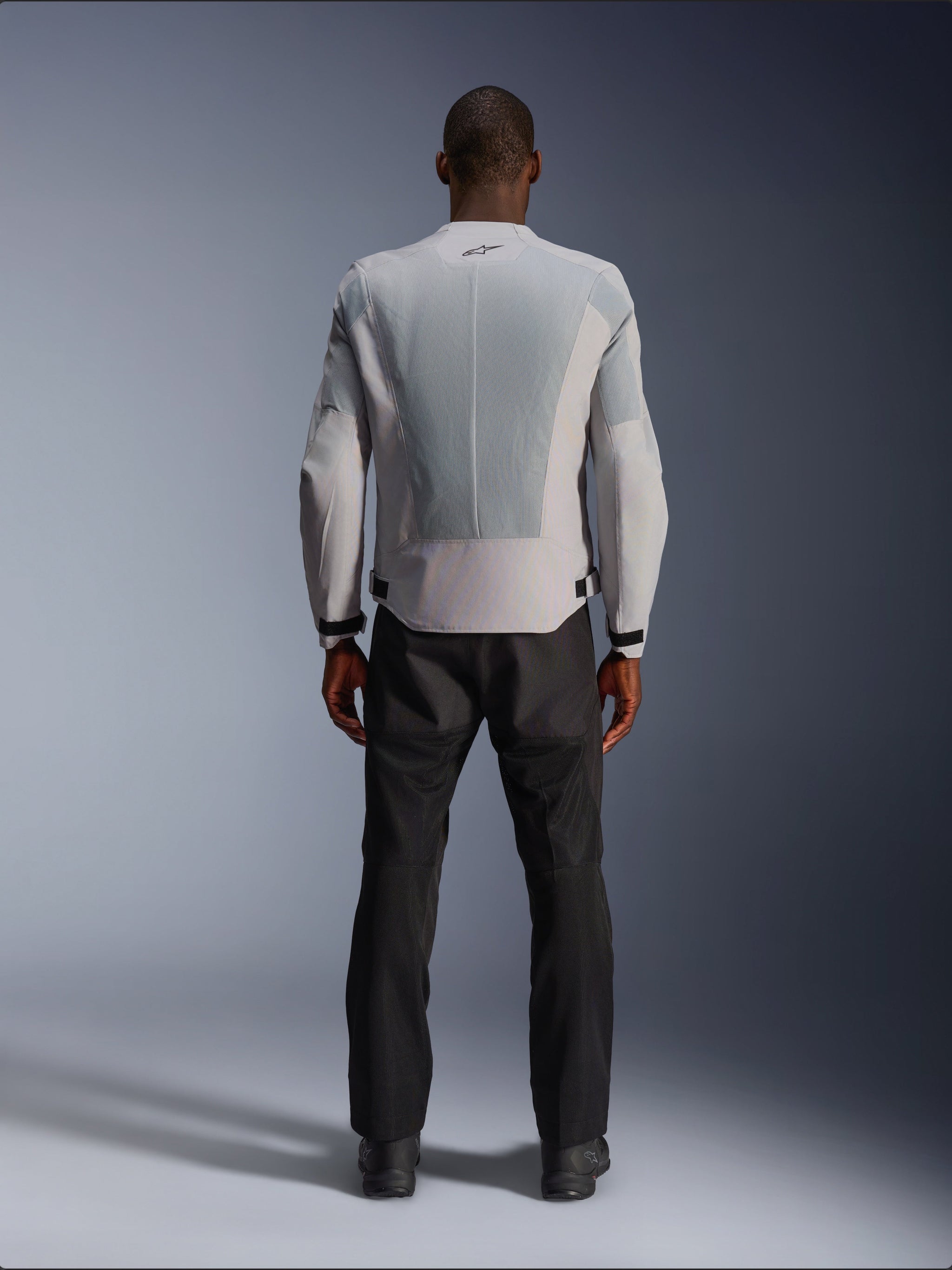 Alpinestars C-1 Air Jacket in Silver, viewed from the back on a male model, textile motorcycle jacket featuring large tech mesh ventilation panels, small black logo at the neck, and waist adjusters, worn with black riding pants for an urban contemporary look.