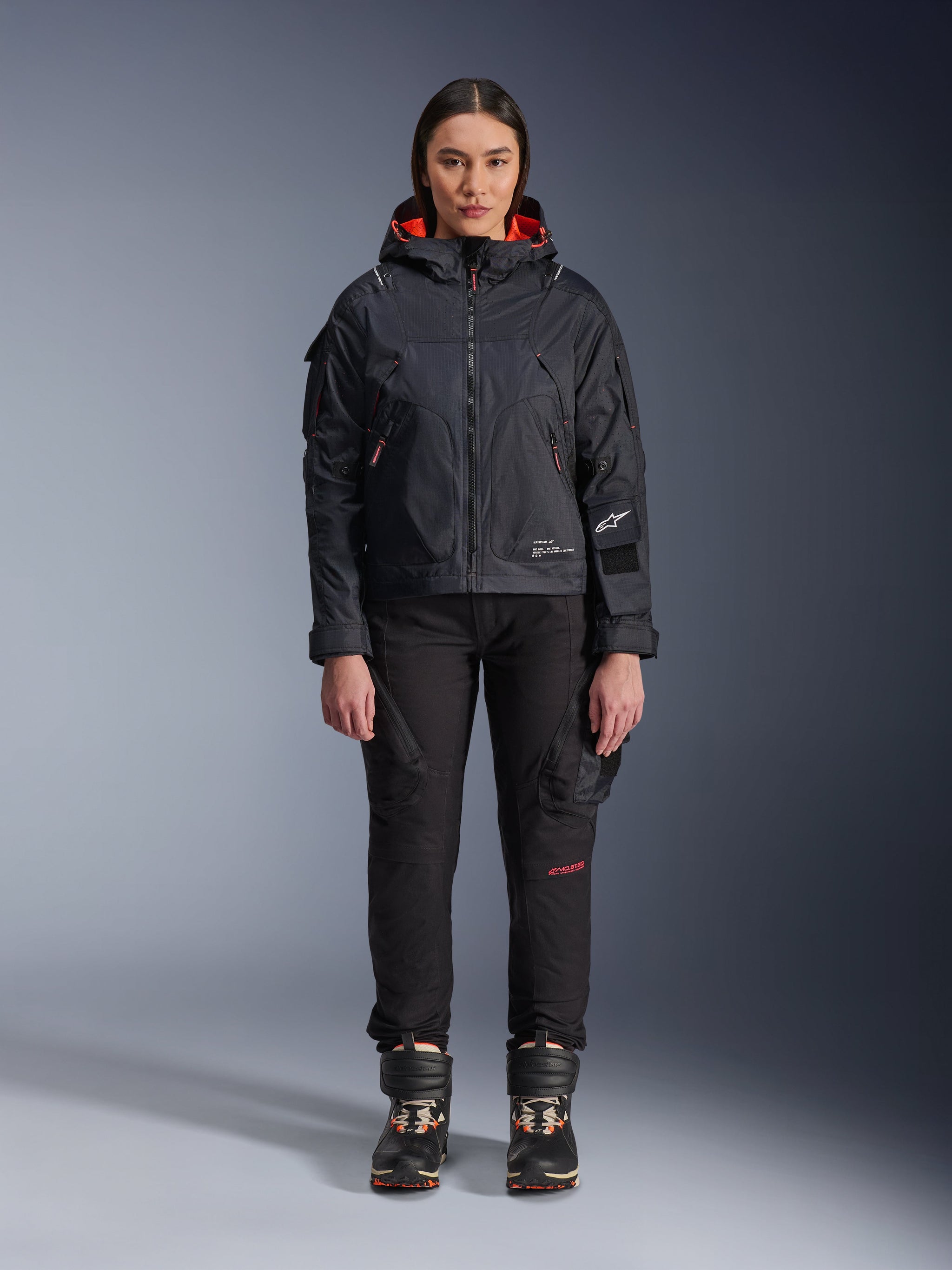 Female model wearing the Alpinestars Molly Air Jacket in Black and Fluo Coral, featuring a utility-inspired design with a hood and coral inner lining, paired with black riding pants and motorcycle boots, urban contemporary motorcycle gear