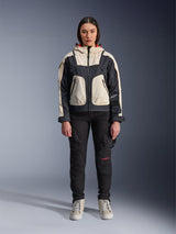 Female model wearing the Alpinestars Molly Air Jacket in Black Almond, featuring a color-blocked design with cream and black panels and a red-lined hood, paired with black motorcycle pants and cream sneakers, studio shot of urban contemporary riding gear.