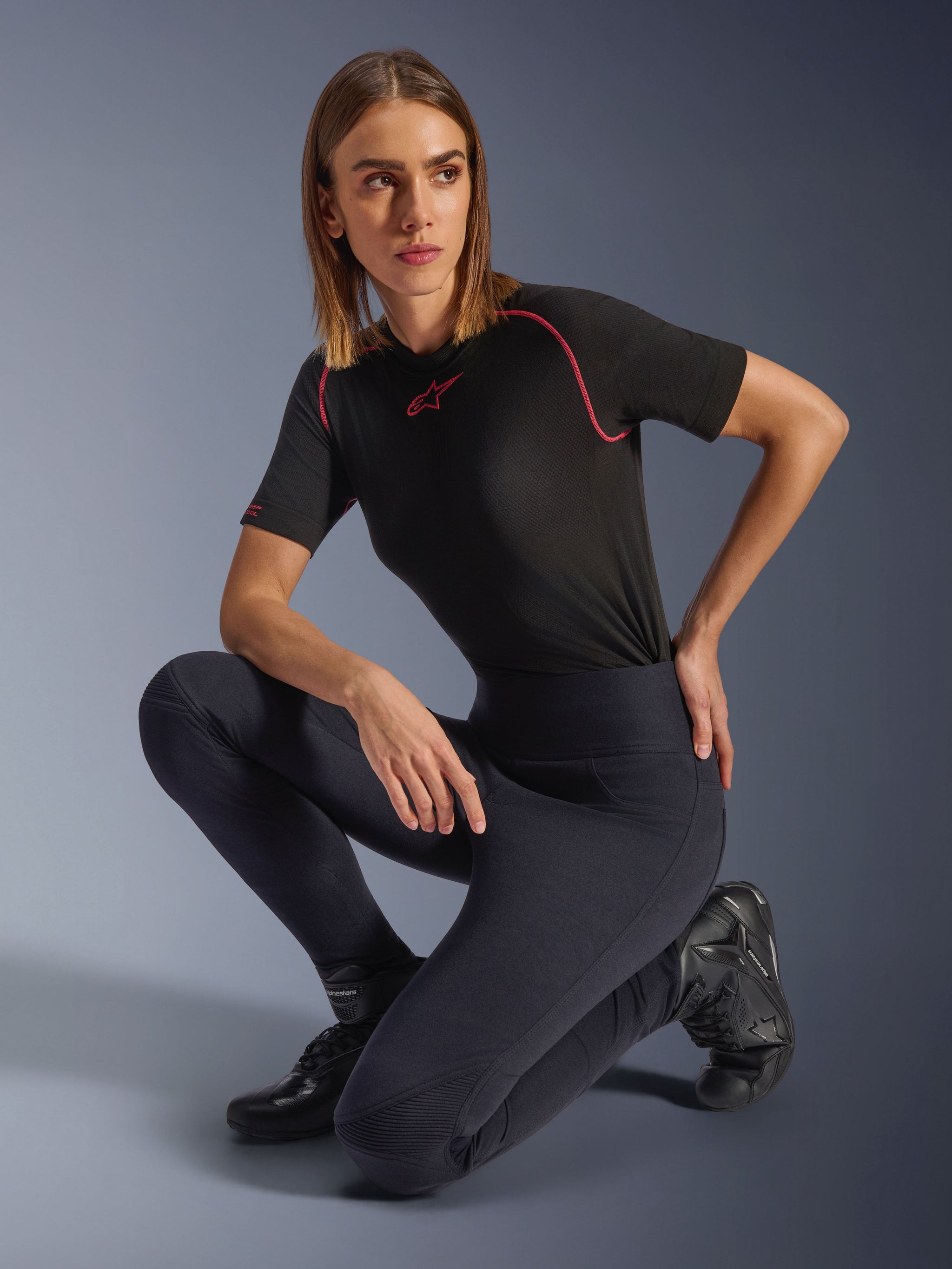 Woman wearing Alpinestars Stella Flex-AST Leggings, Black high-waisted skinny motorcycle leggings with accordion knee panels, styled with a black short-sleeve performance tee with pink stitching and riding boots.