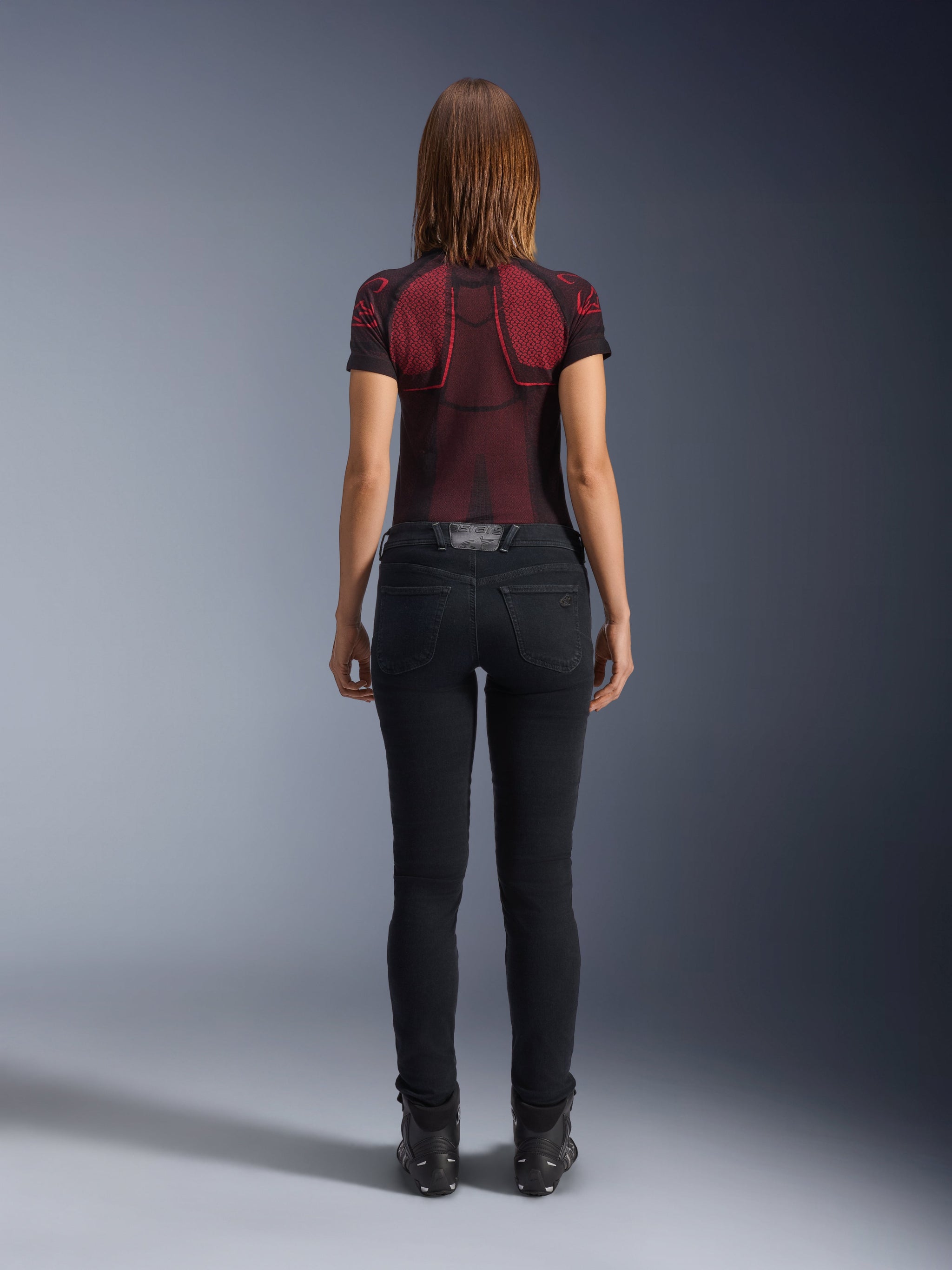 Alpinestars Daisy V3 Women's Riding Denim, Rinse Black, slim fit motorcycle pants shown from behind, featuring a high-waist design and back pockets, paired with a red and black patterned technical shirt and black boots, urban motorcycle gear for women