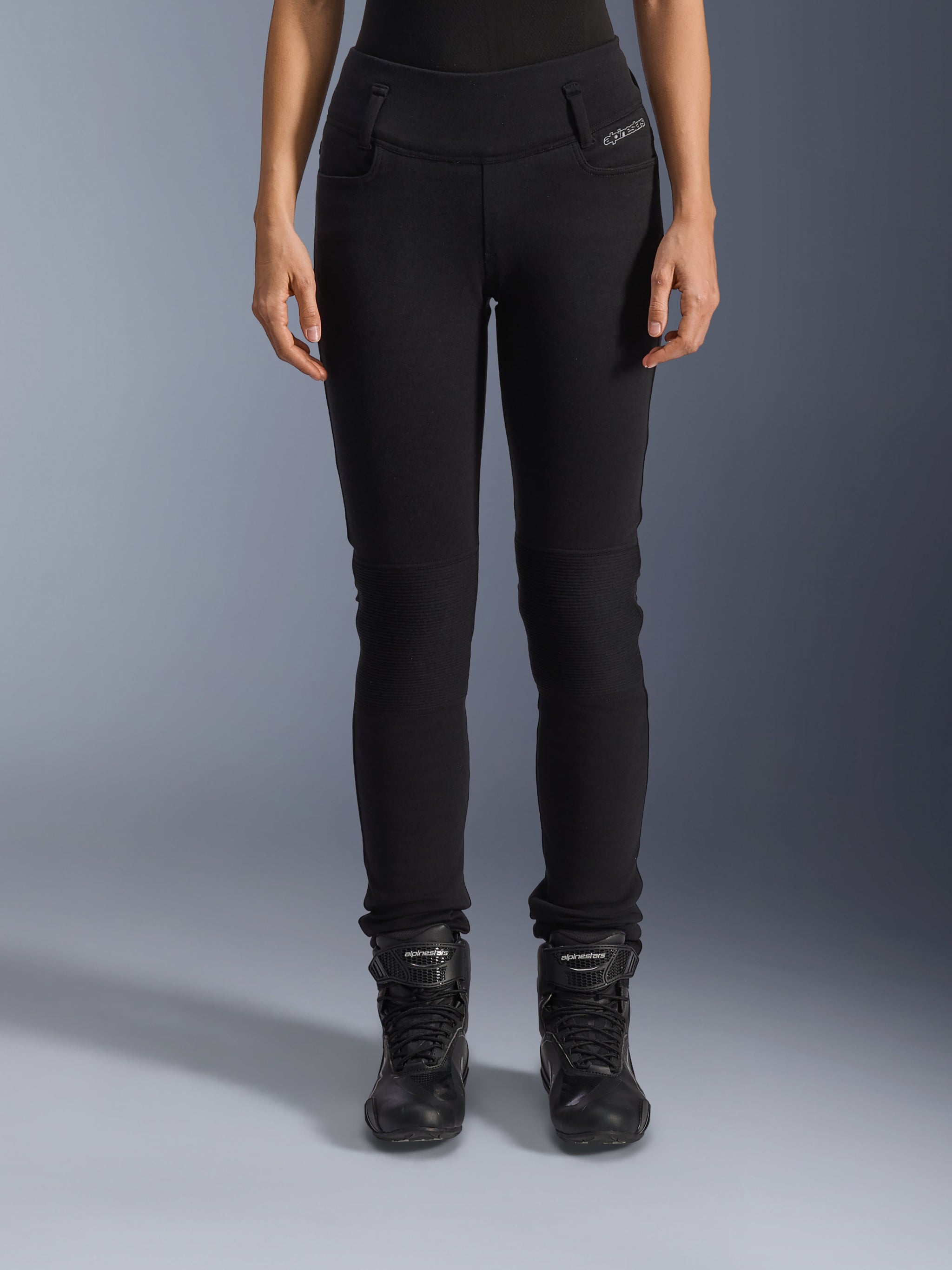 Alpinestars Womens Banshee Leggings, Black motorcycle pants, slim-fit design with accordion knee panels, worn with black high-top riding boots, urban road gear with a casual style