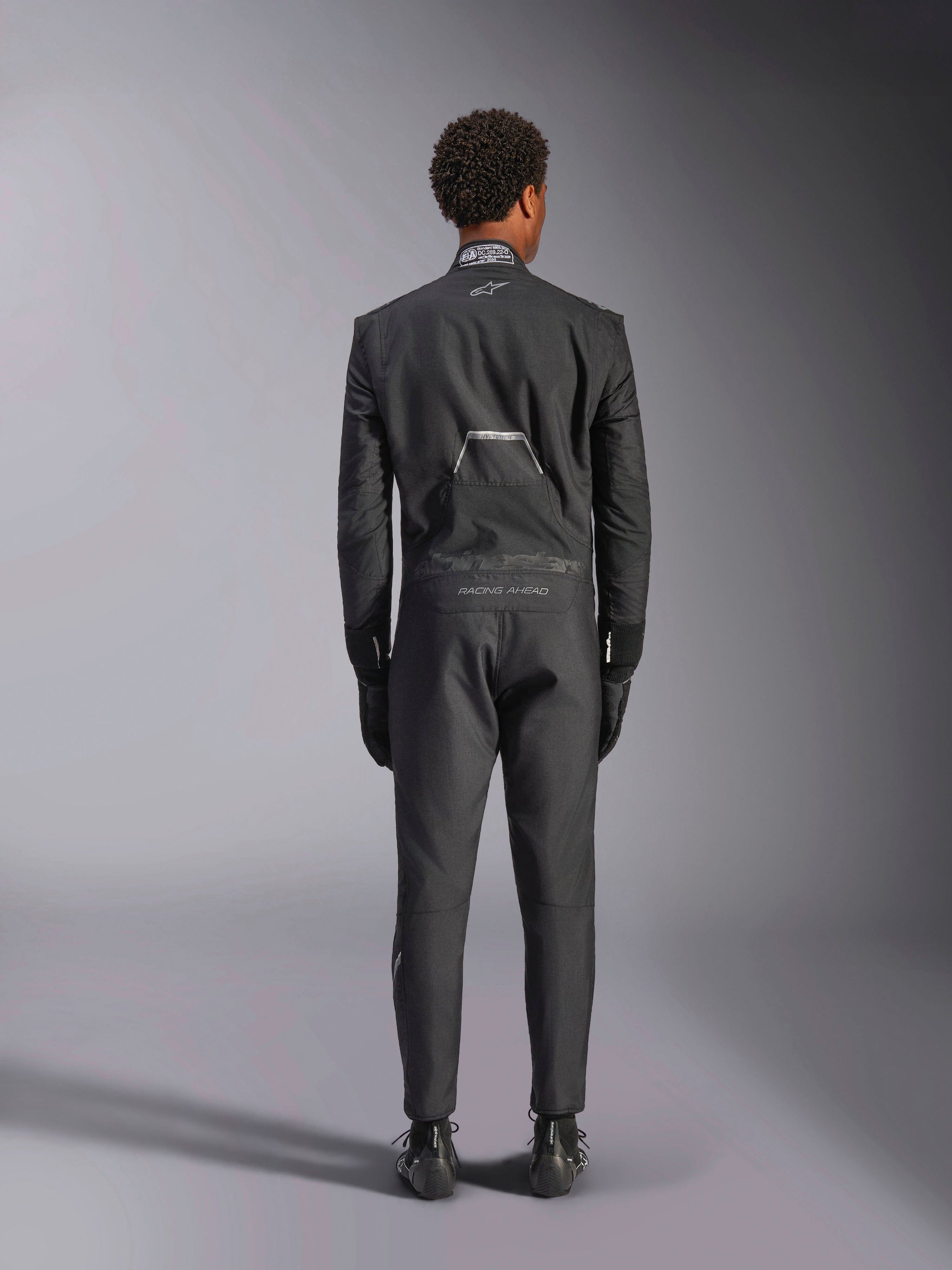 Alpinestars Hypertech V3 Suit, Black Dark Shadow, rear view of a professional one-piece auto racing suit, featuring FIA certification label on the collar and 'Alpinestars' and 'RACING AHEAD' branding on the lower back, worn with matching black racing gloves and boots.
