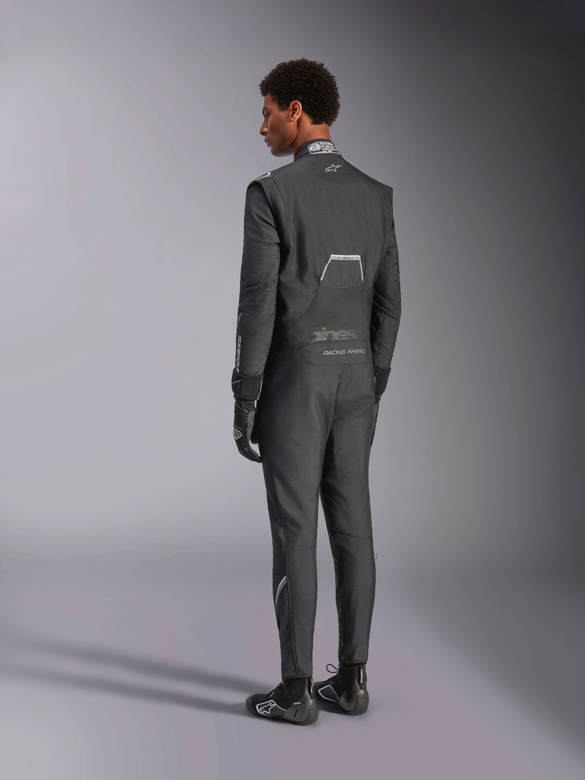 Alpinestars Hypertech V3 Suit, Black and Dark Shadow, rear view of male model wearing professional auto racing suit with matching black gloves and boots, featuring lightweight Nomex construction and FIA certification on the collar, athletic-slim fit for motorsport.