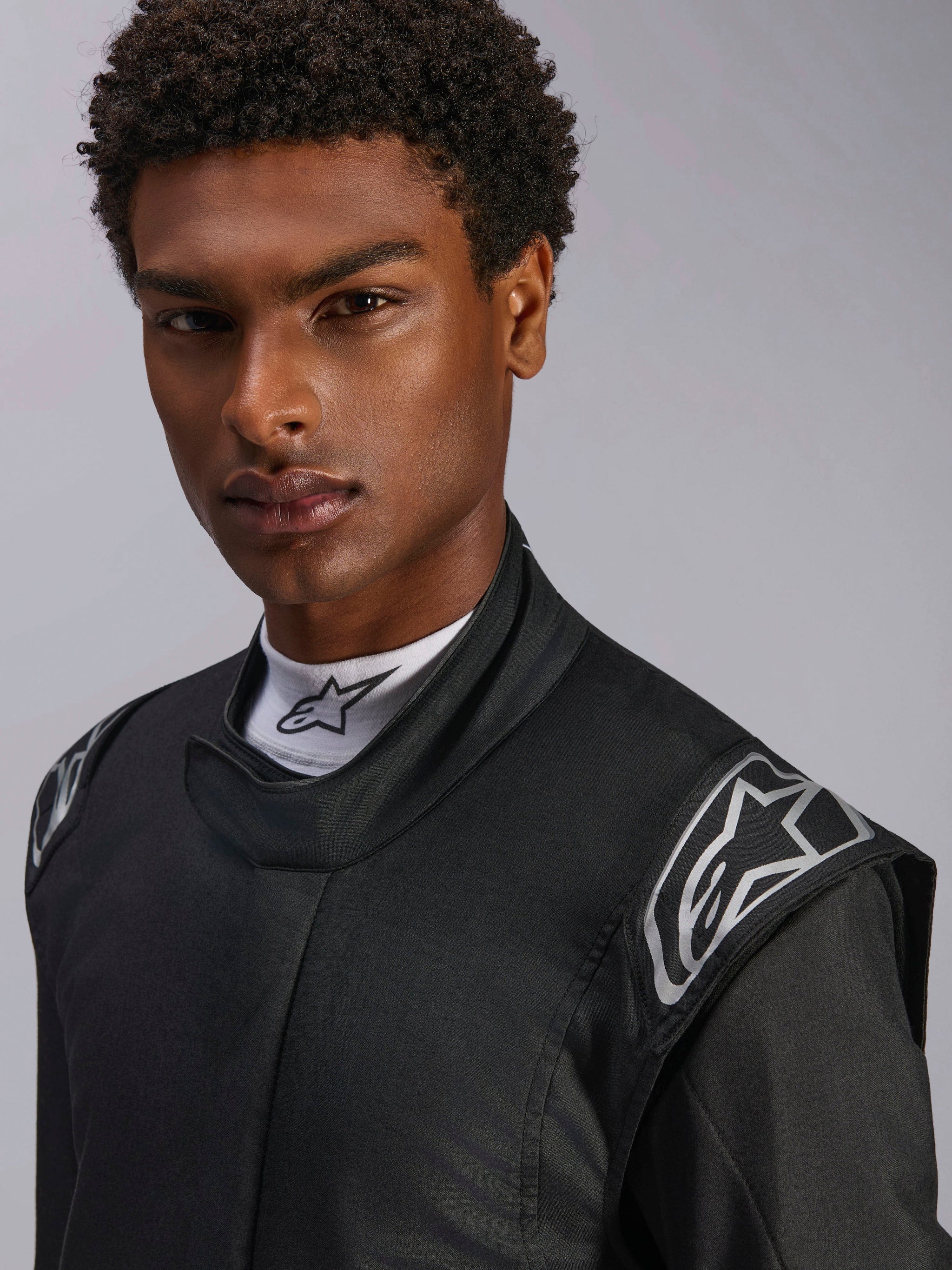 Alpinestars Hypertech V3 Suit, Black Dark Shadow auto racing suit, close-up on male model, silver shoulder logos, high collar with white Alpinestars mock neck shirt, professional motorsport protective gear